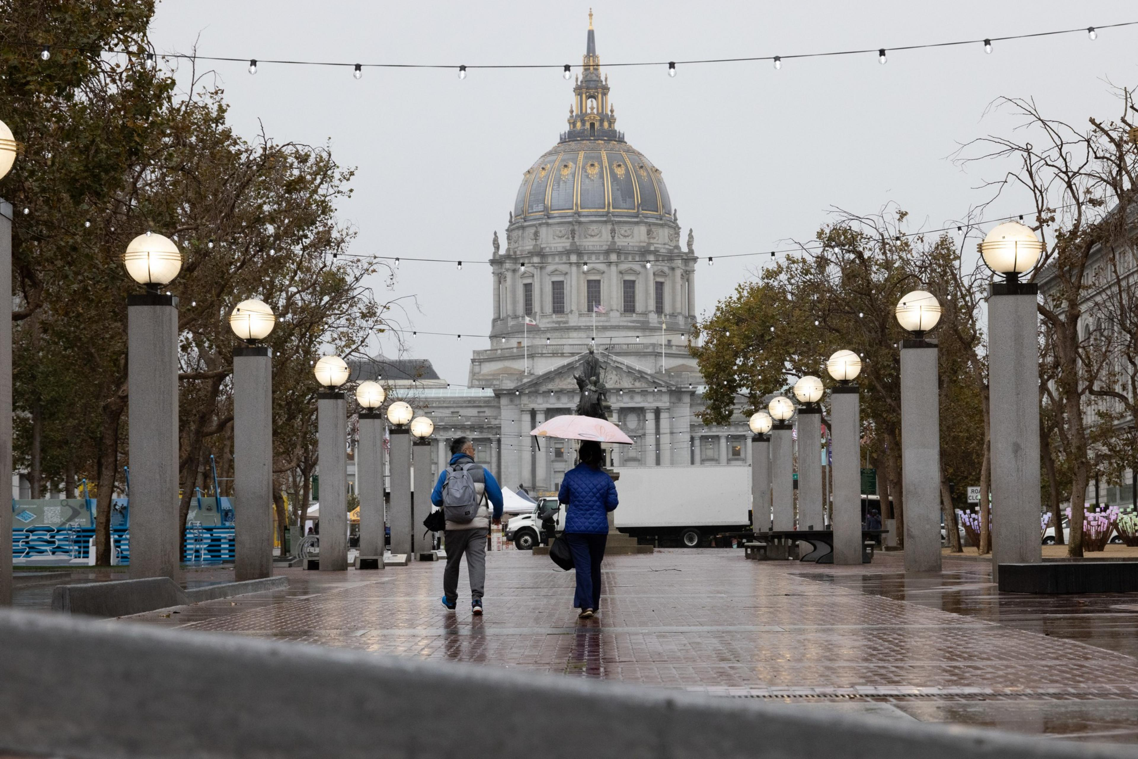 Two people walk down a wet pathway flanked by trees and light posts, with a large domed building in the background under a gray sky.