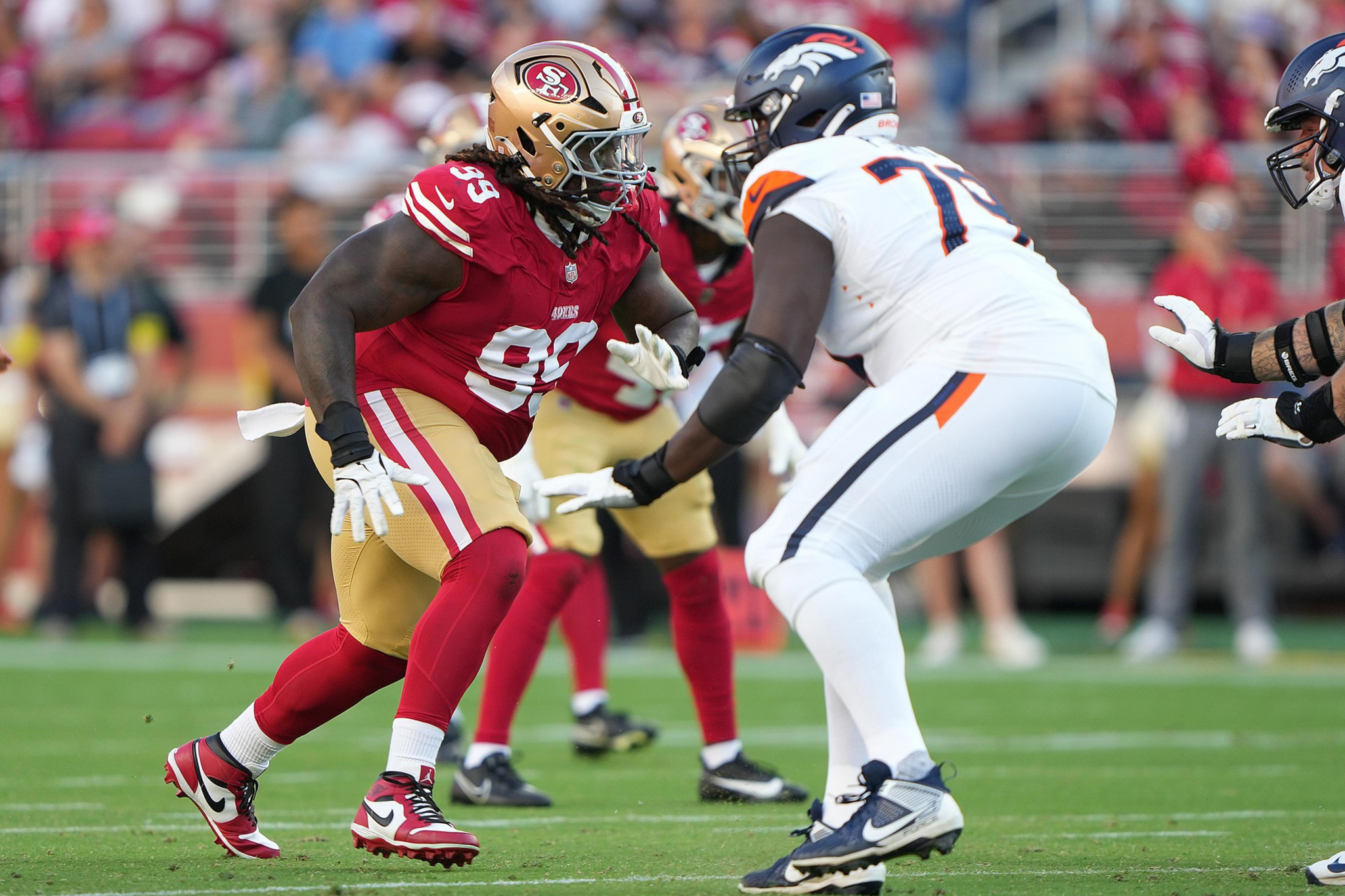 A San Francisco 49ers player in red and gold faces off against a Denver Broncos player in white and navy during an intense football game moment.