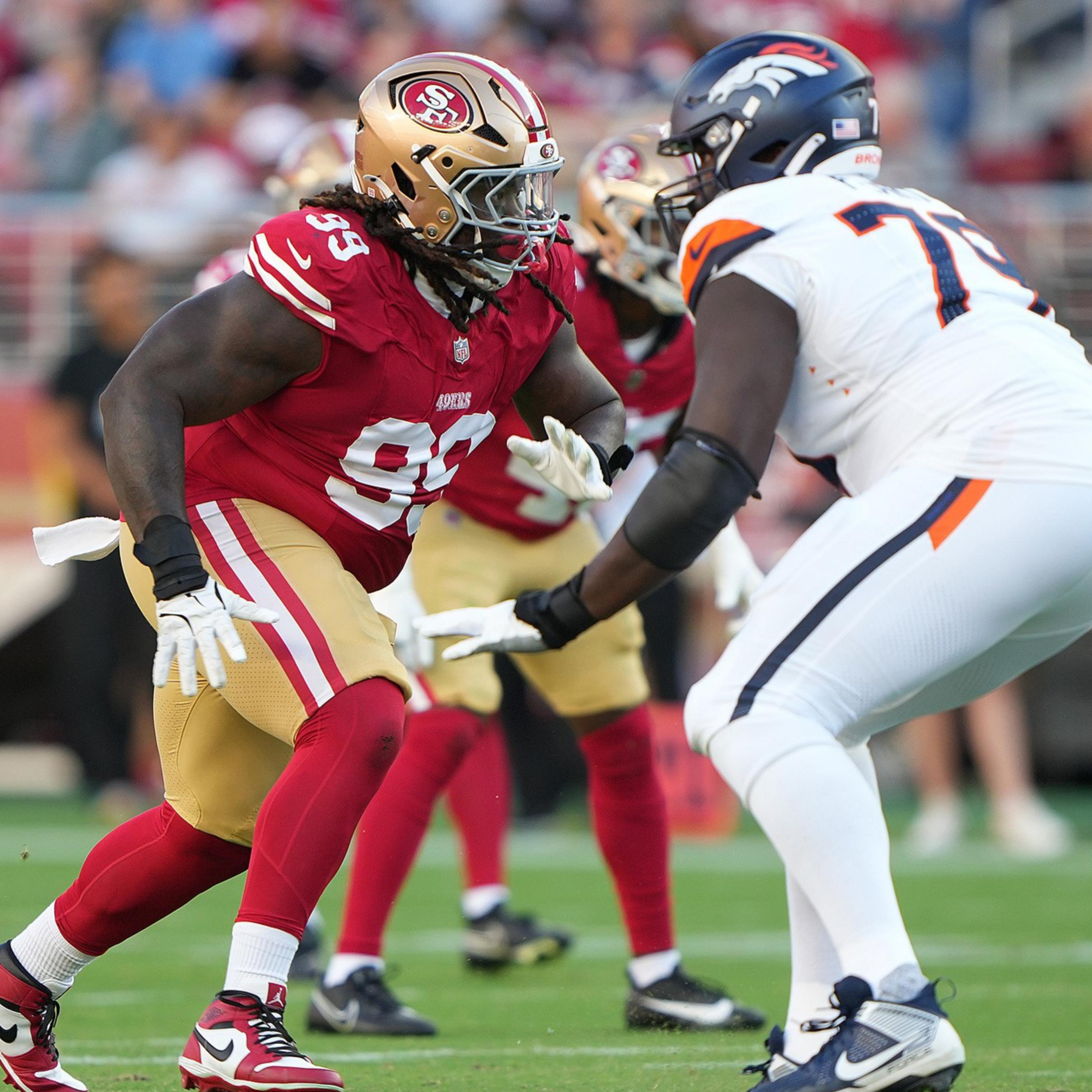 A San Francisco 49ers player in red and gold faces off against a Denver Broncos player in white and navy during an intense football game moment.