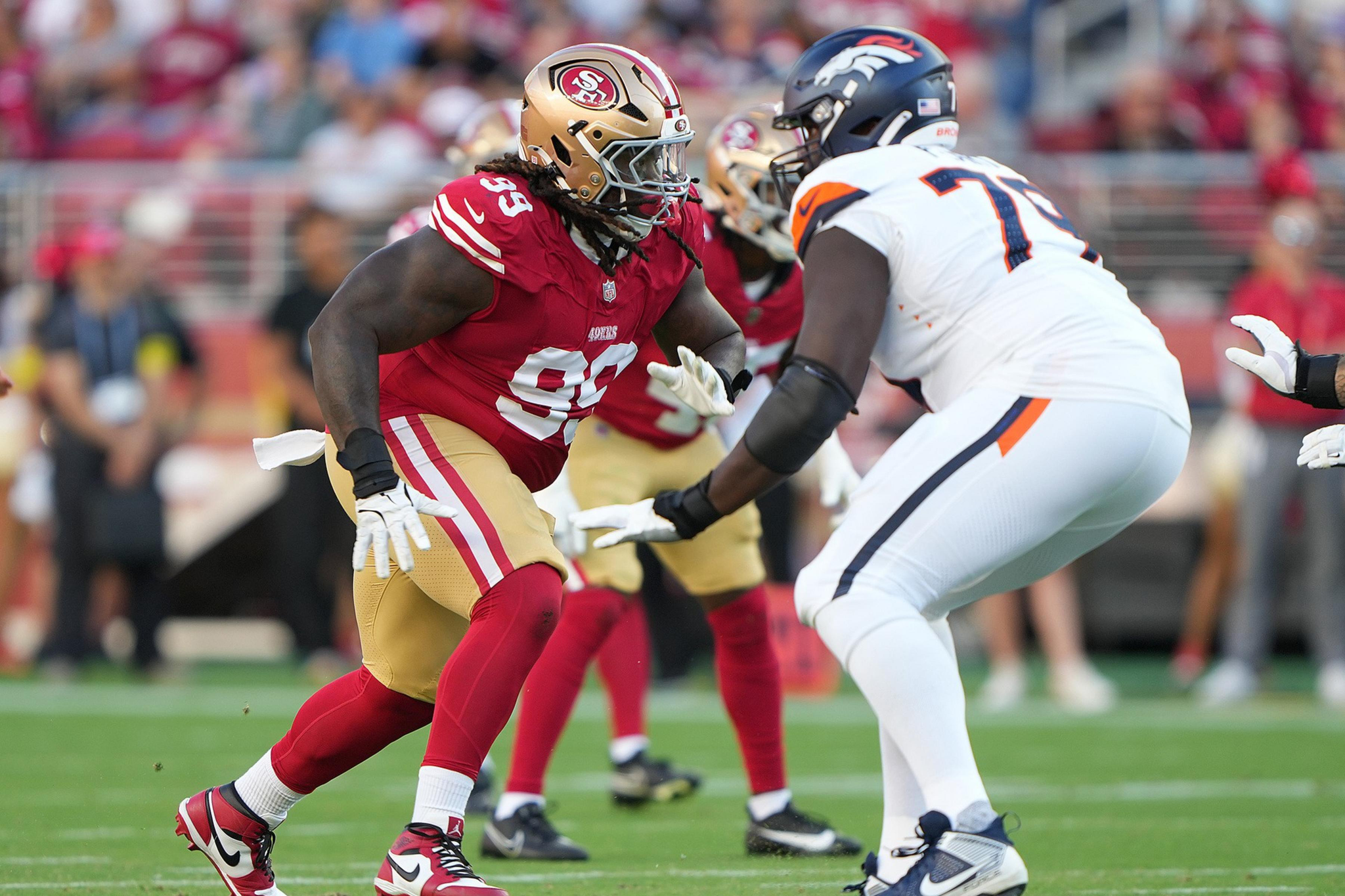 A San Francisco 49ers player in red and gold faces off against a Denver Broncos player in white and navy during an intense football game moment.