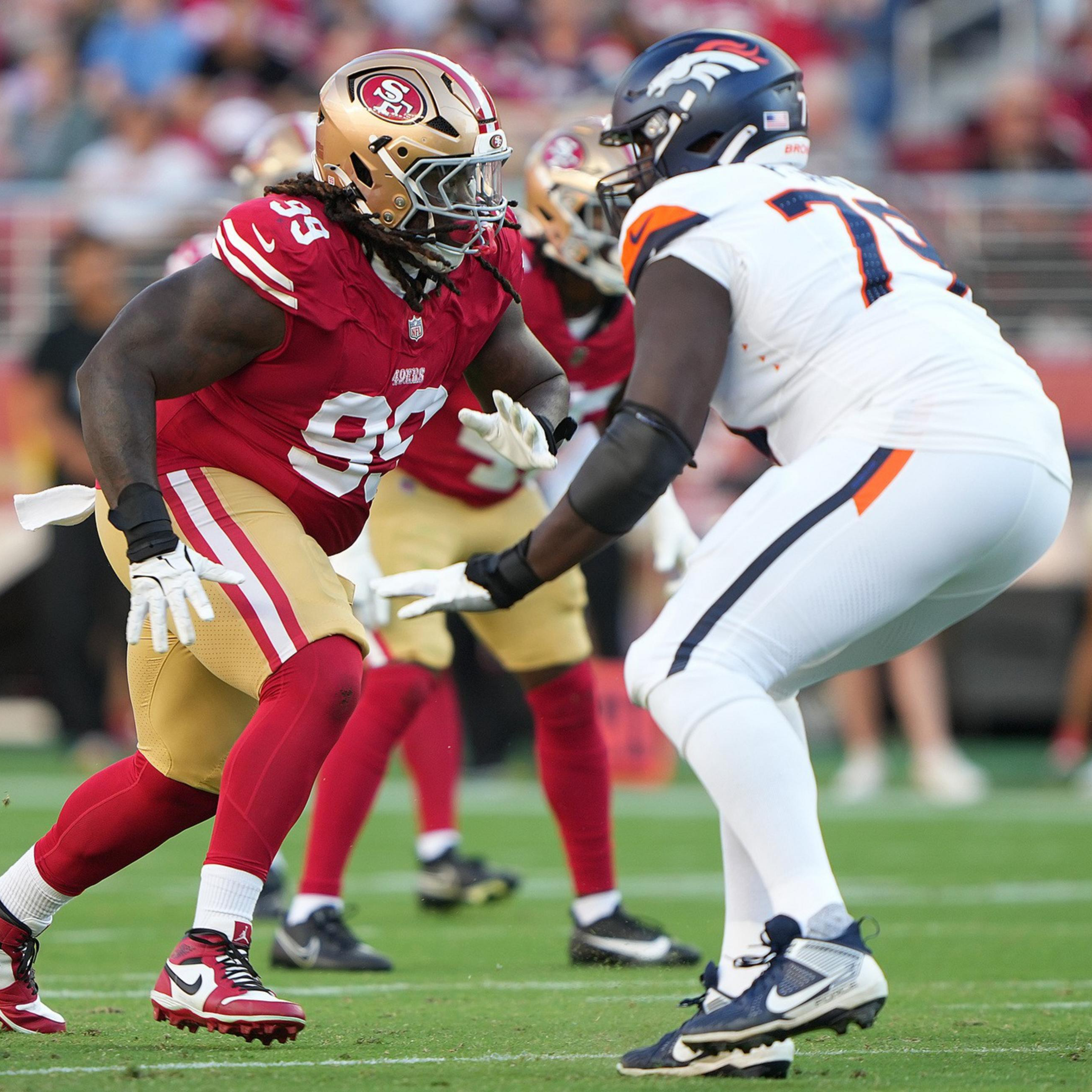 A San Francisco 49ers player in red and gold faces off against a Denver Broncos player in white and navy during an intense football game moment.