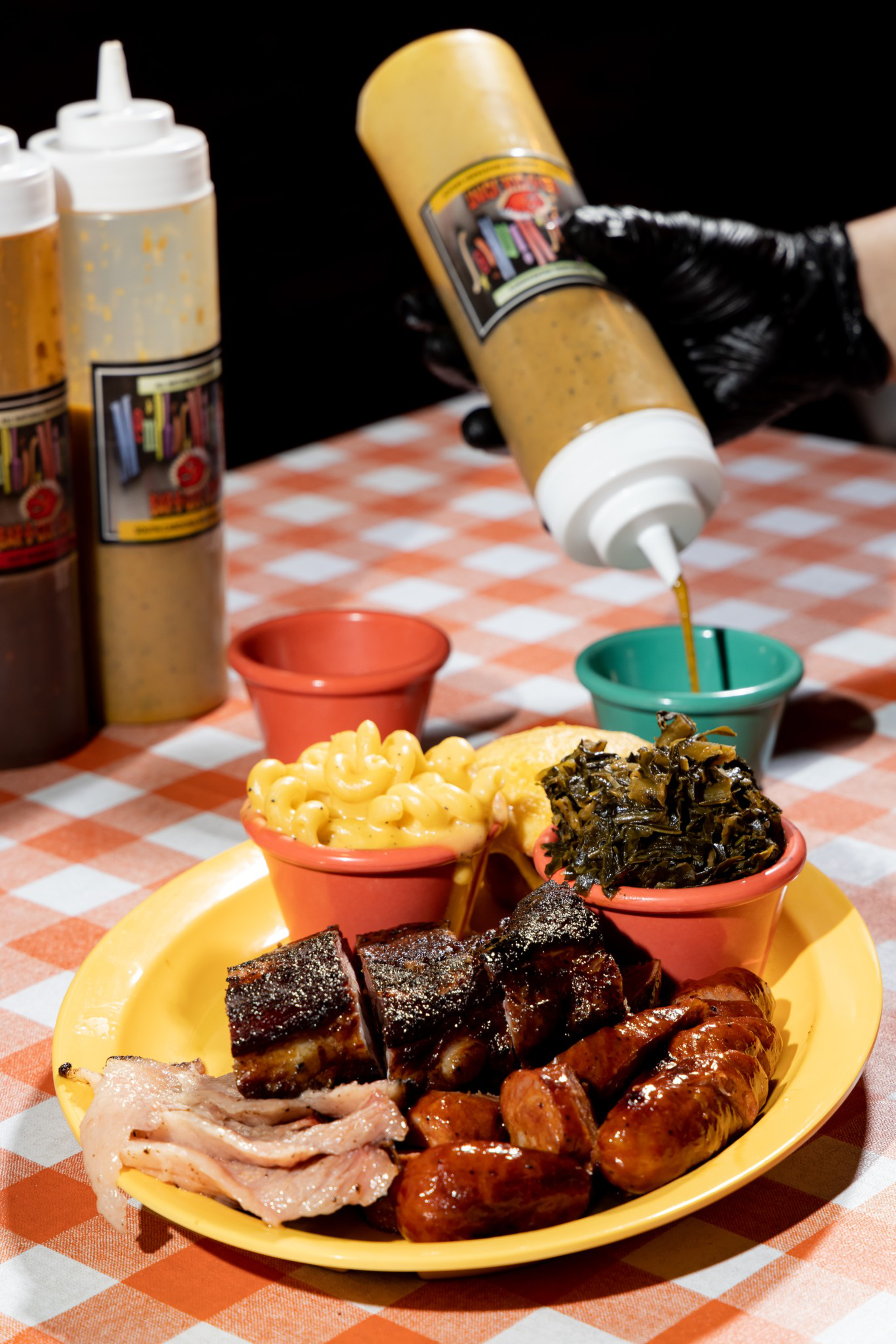 Barbecue plate with ribs, sausage, bacon, mac and cheese, and greens served in colorful cups; sauce is being poured from a squeeze bottle.