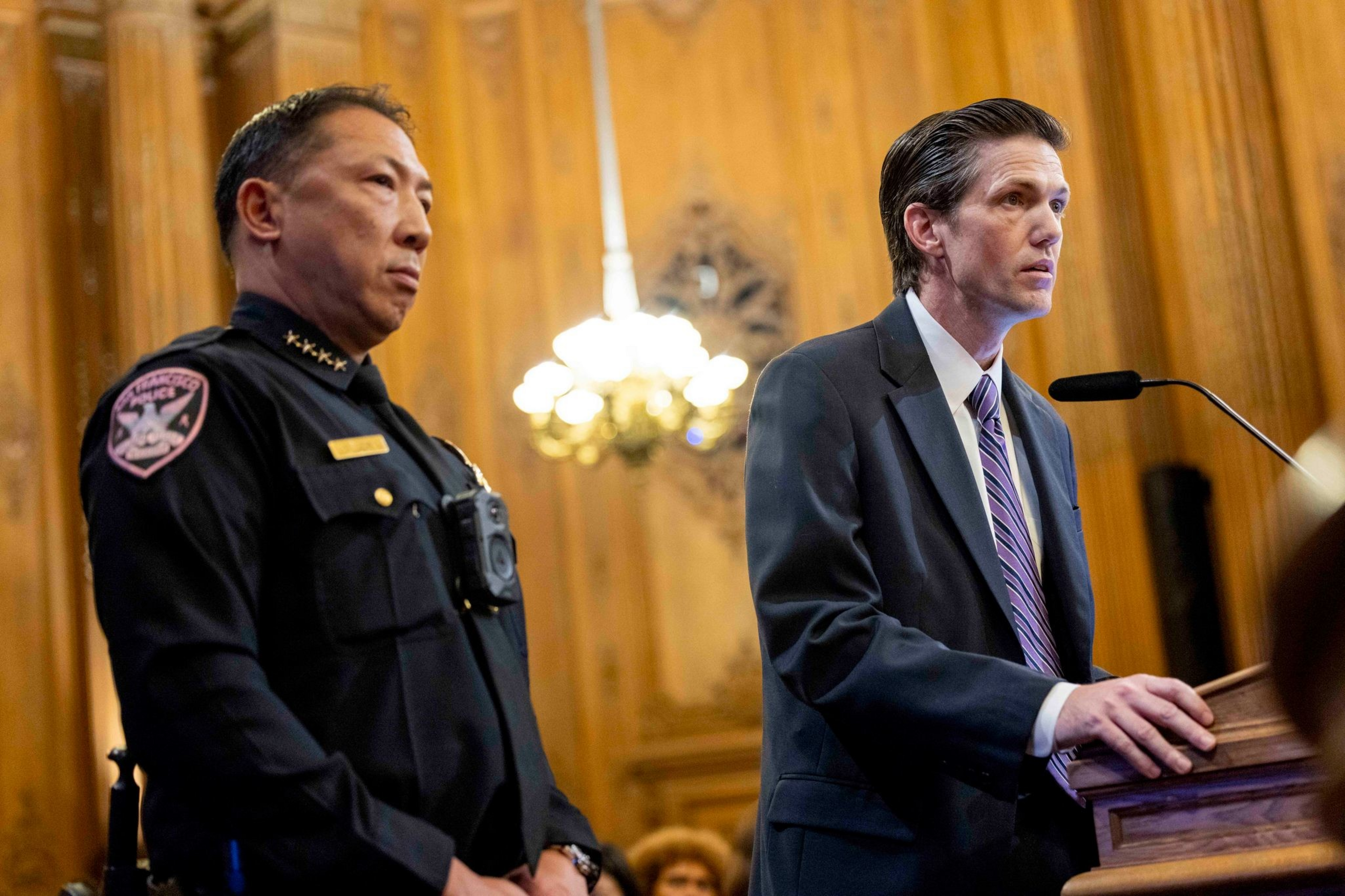 A uniformed police officer stands next to a man in a suit speaking at a wooden podium with a microphone in a wood-paneled room.