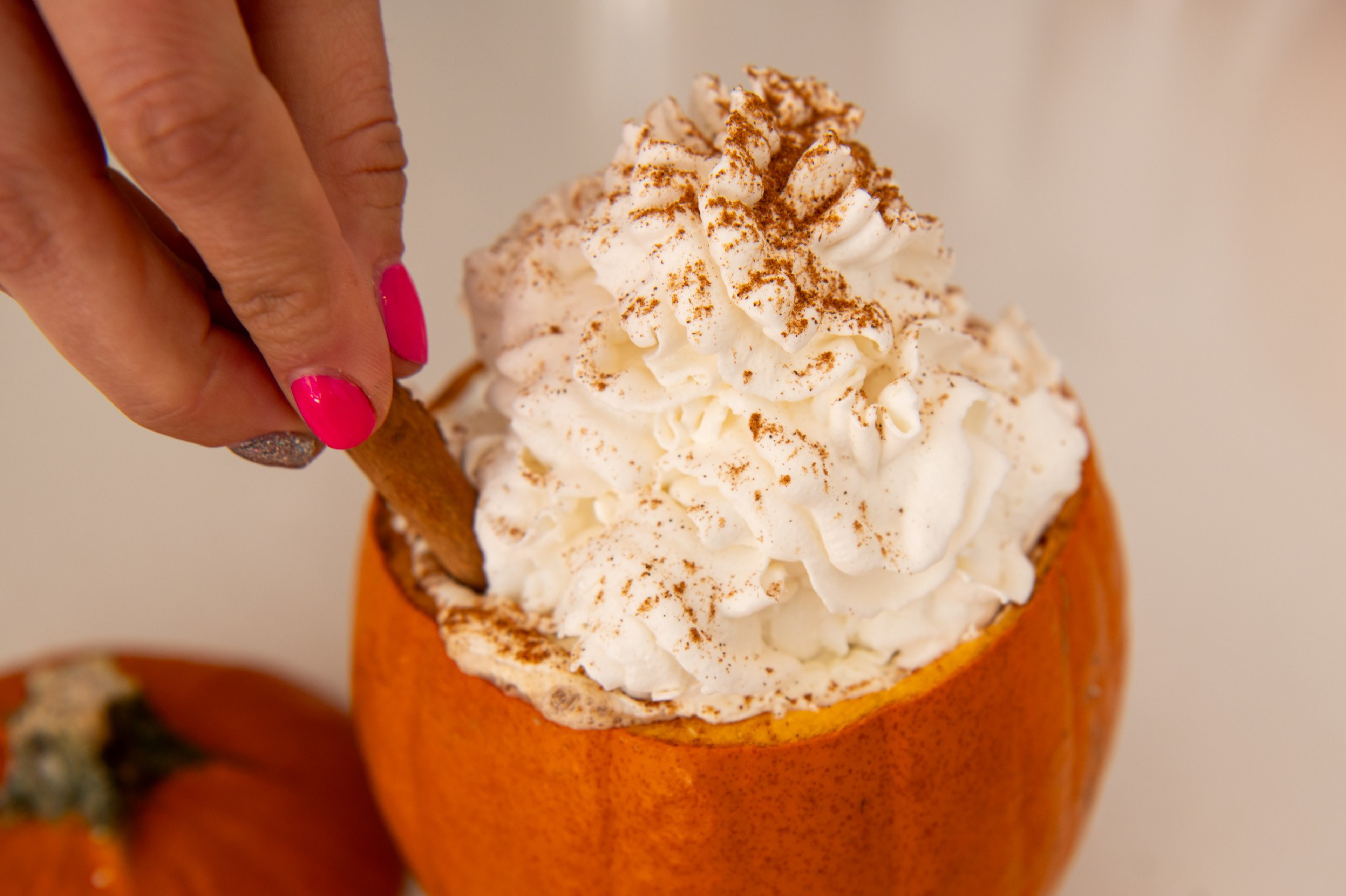 A hand with pink and silver nail polish is dipping a cinnamon stick into a small pumpkin filled with whipped cream topped with cinnamon powder.