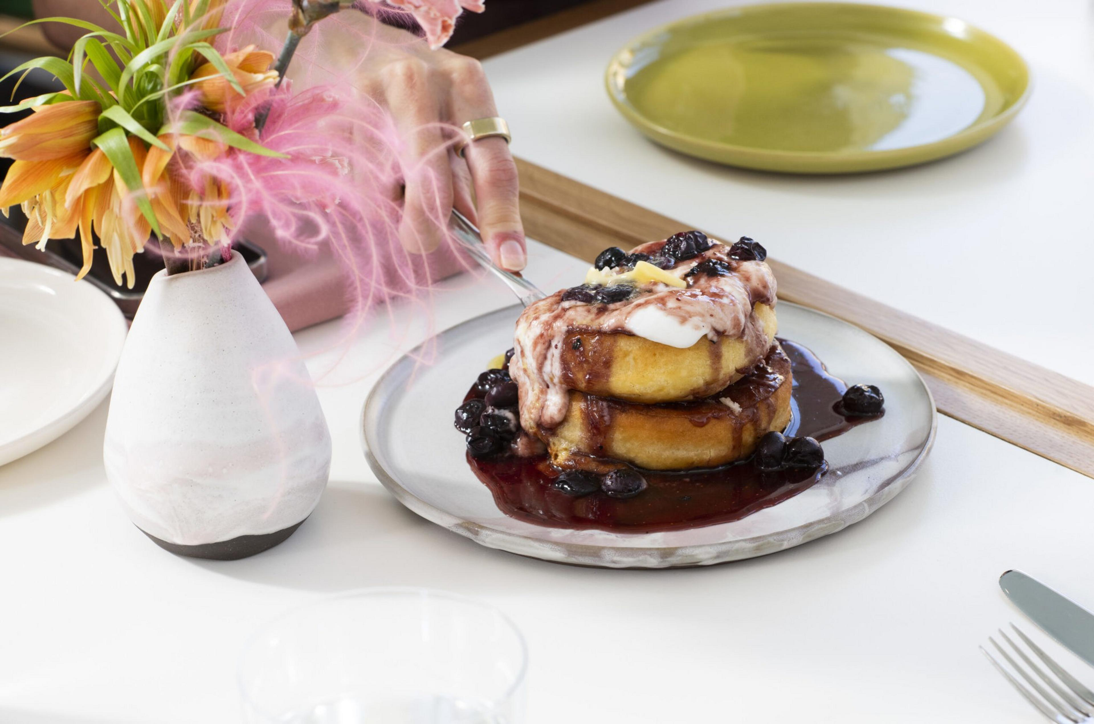 Two pancakes stacked with blueberry sauce, butter, and cream sit on a plate, while a hand holds a fork nearby and a flower vase is on the table.