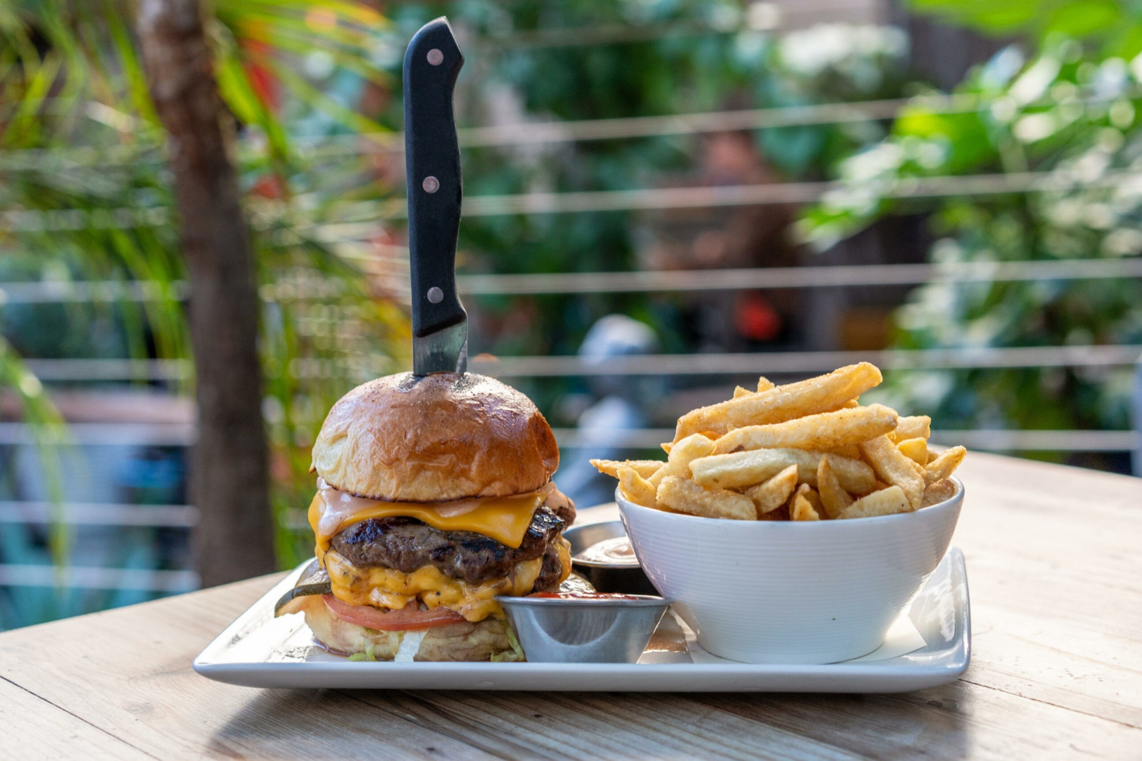 A cheeseburger with a knife stuck in its bun is served on a white plate alongside a bowl of golden French fries and dipping sauces.