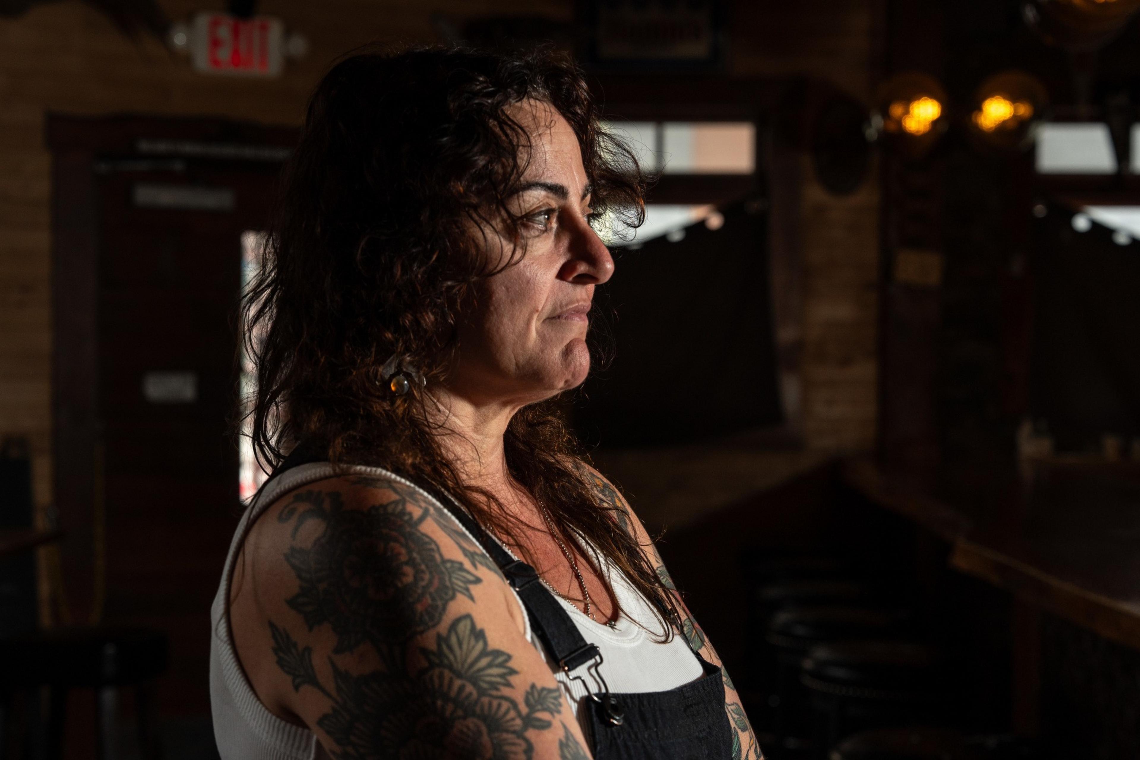 A woman with curly hair and extensive arm tattoos is gazing to the side in a dimly lit interior. She wears a white shirt and black overalls.