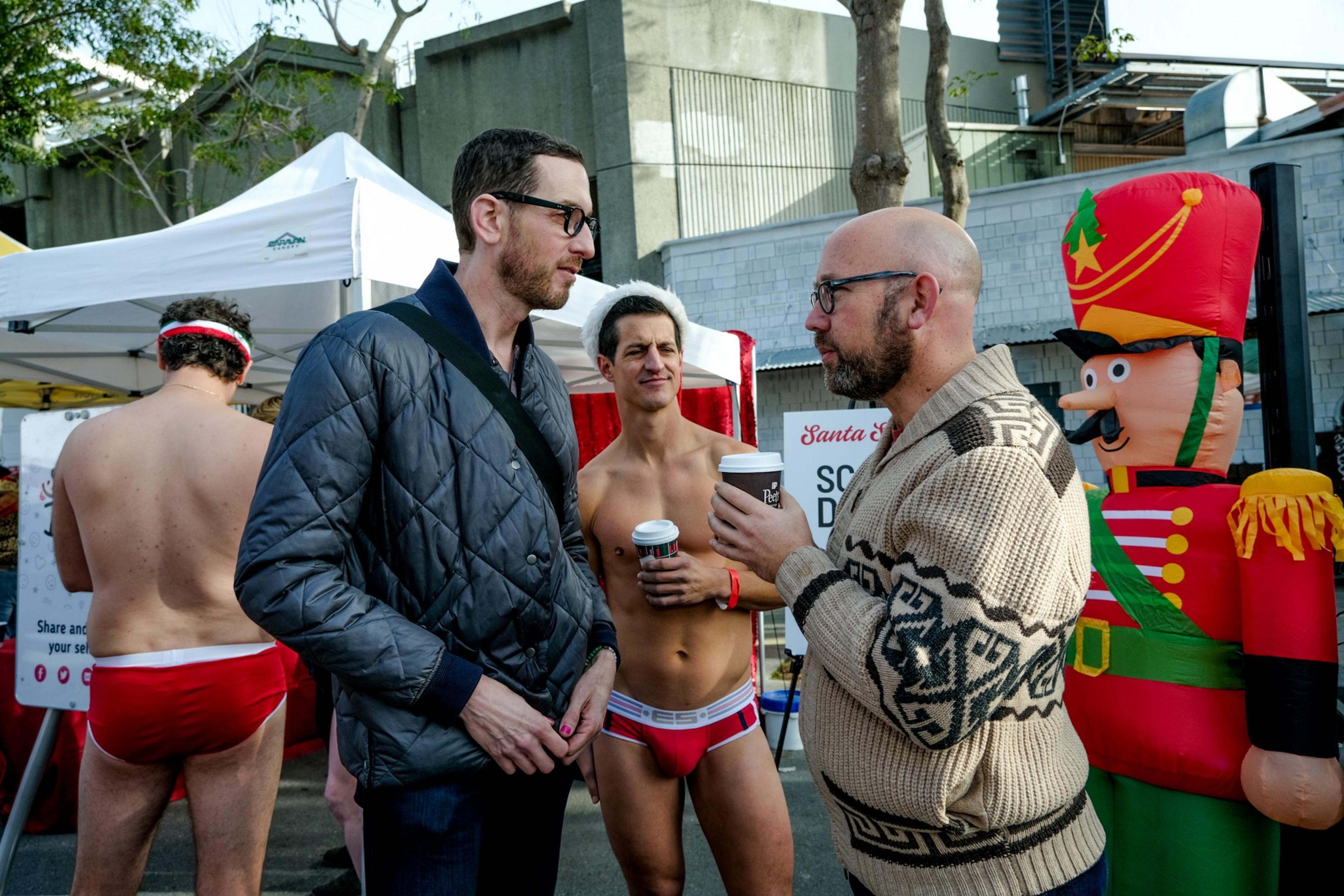 Two men wearing glasses talk to each other as a shirtless man in a Santa hat looks on