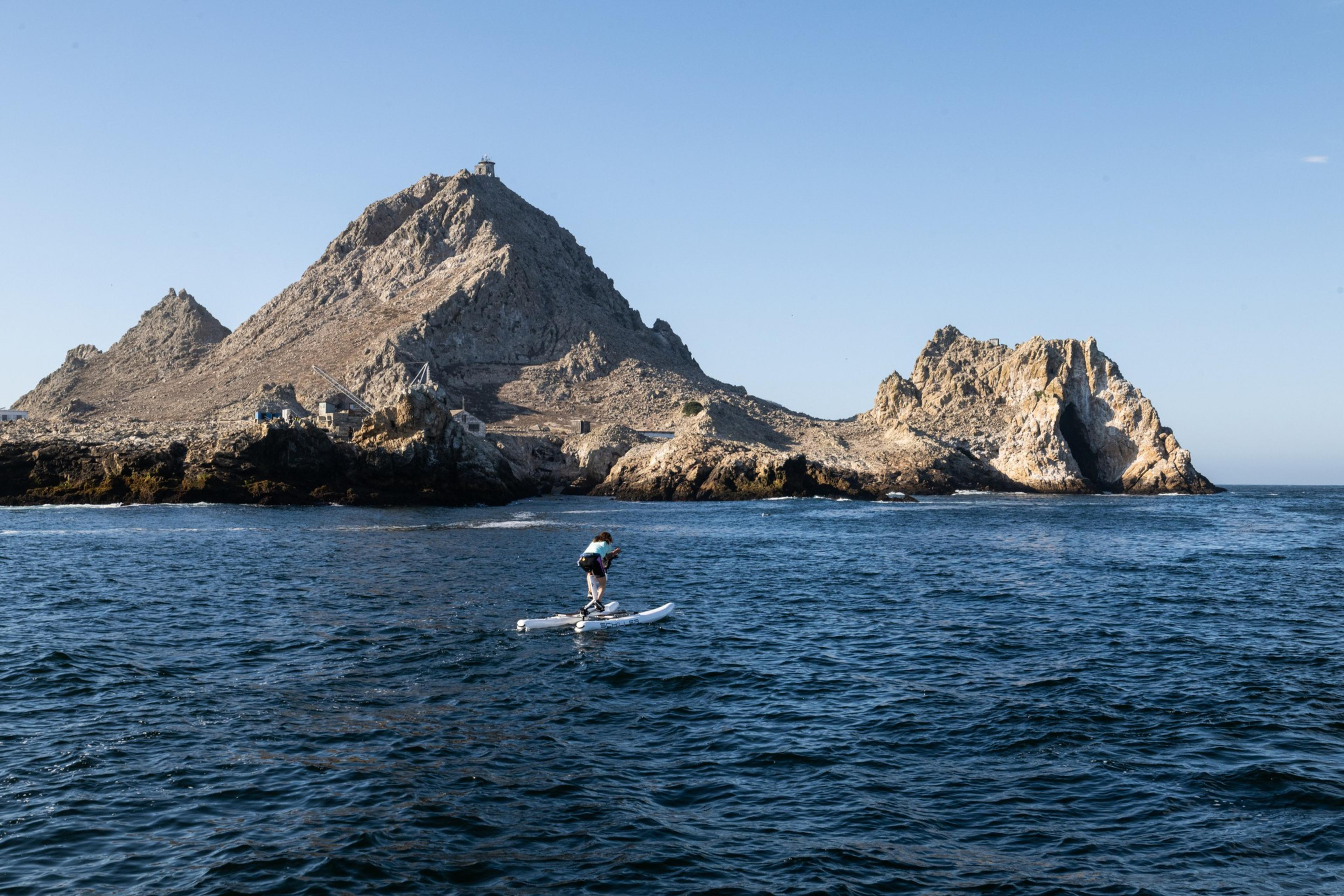 A person rides a pedal-powered watercraft on the blue sea, with rocky islands in the background under a clear blue sky.