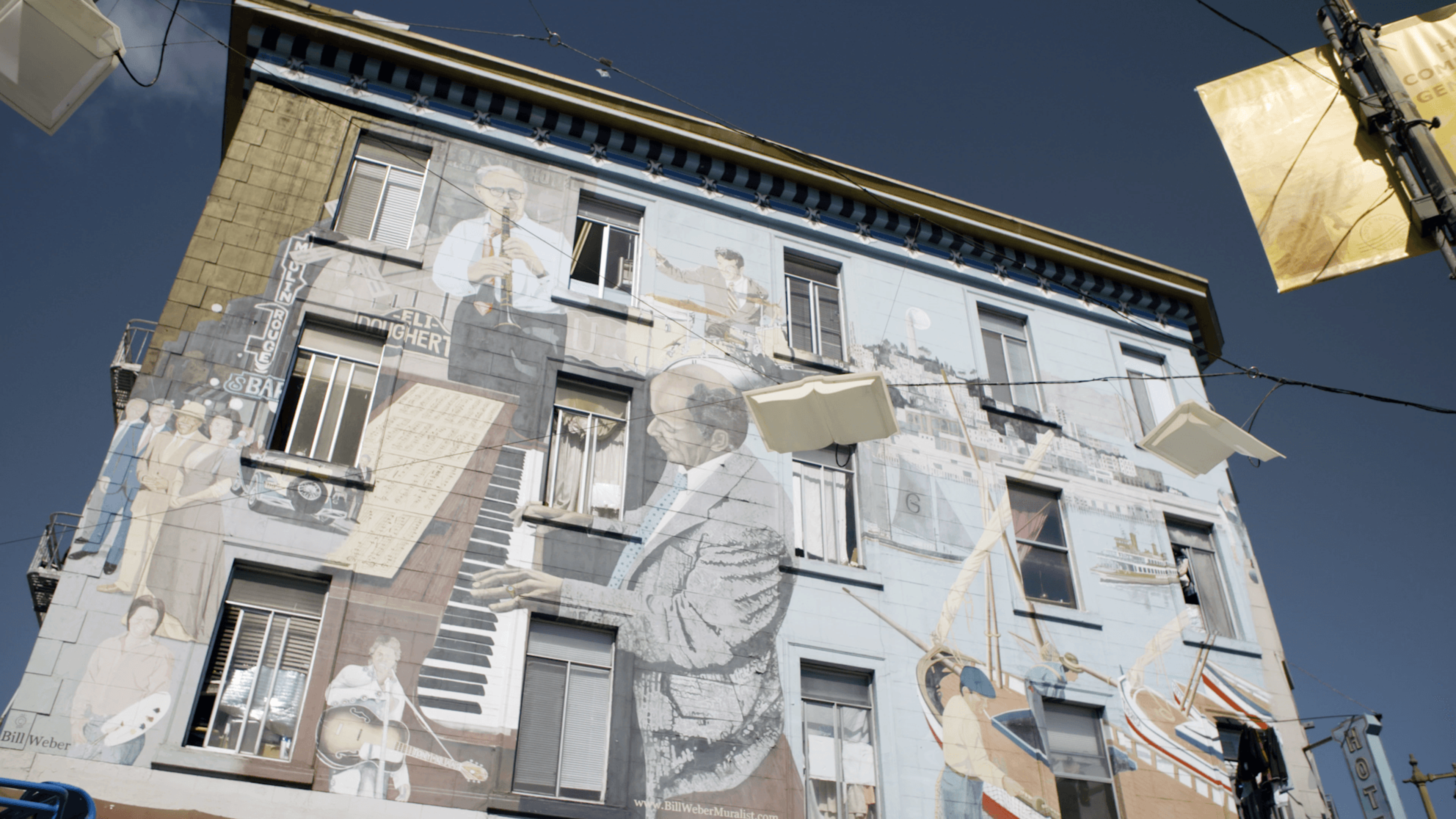 A building wall painted with a mural of musicians playing instruments, people interacting, and a large piano keyboard, under a clear blue sky.