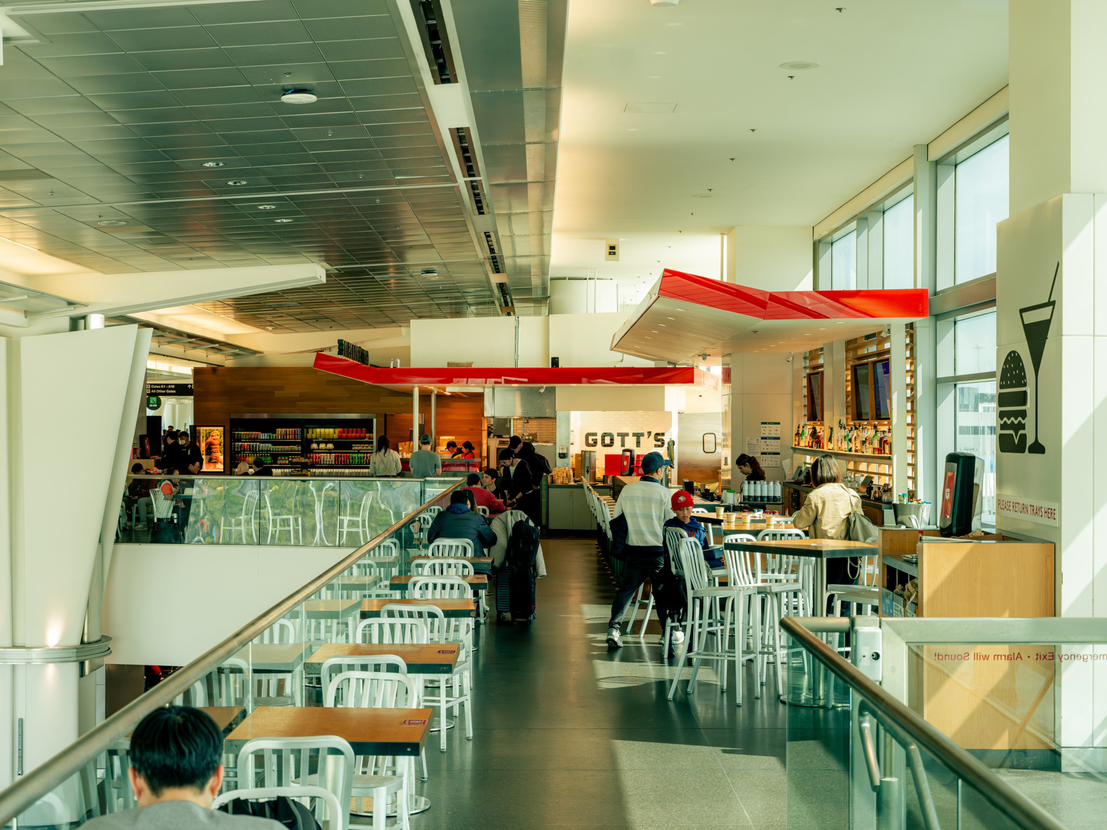 A bright, modern food court with white tables and chairs, people dining, and a red-accented Gott’s restaurant counter at the far end.