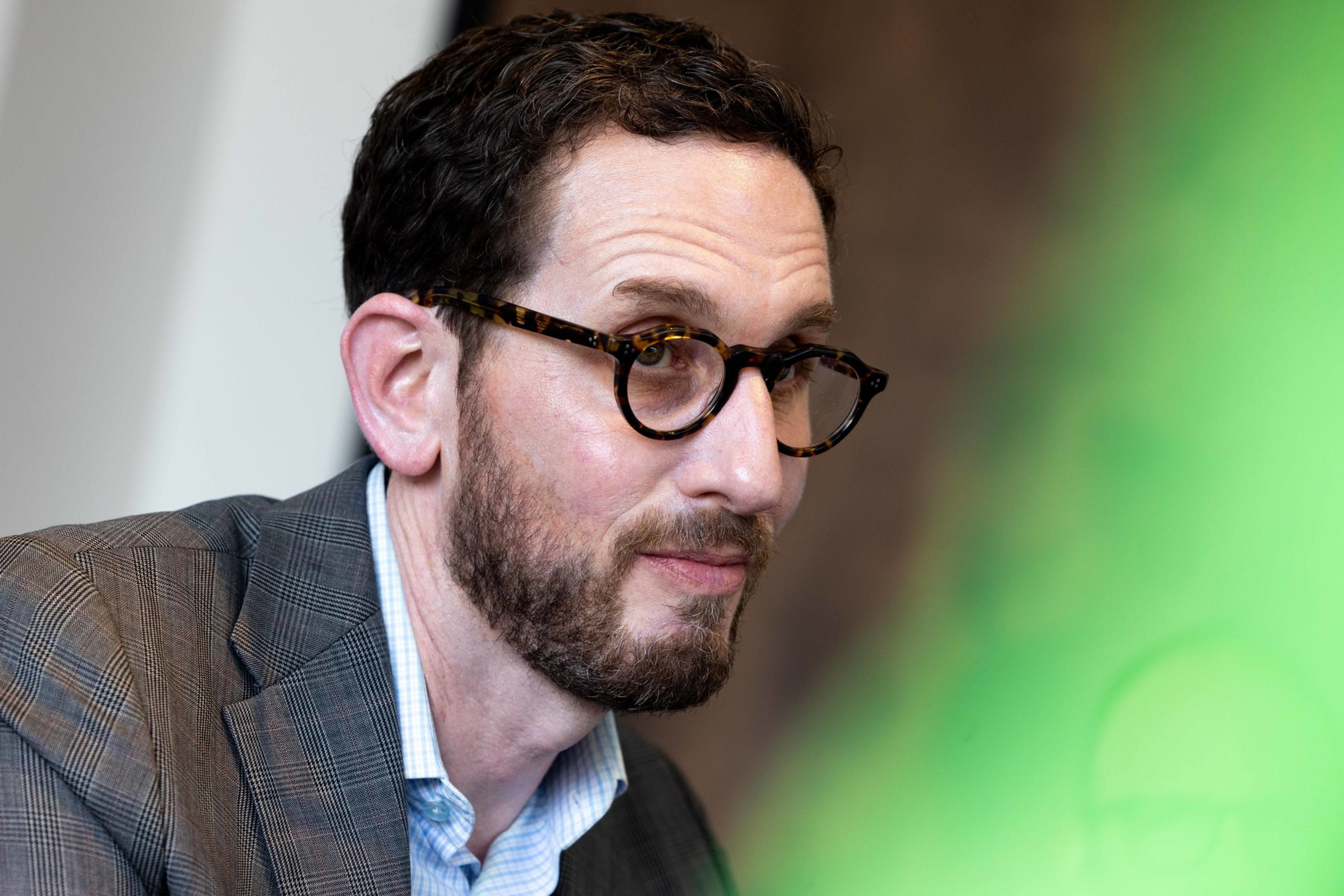 A man with dark hair and a beard wears tortoiseshell glasses, a blue checkered shirt, and a gray plaid blazer, looking intently to the side.