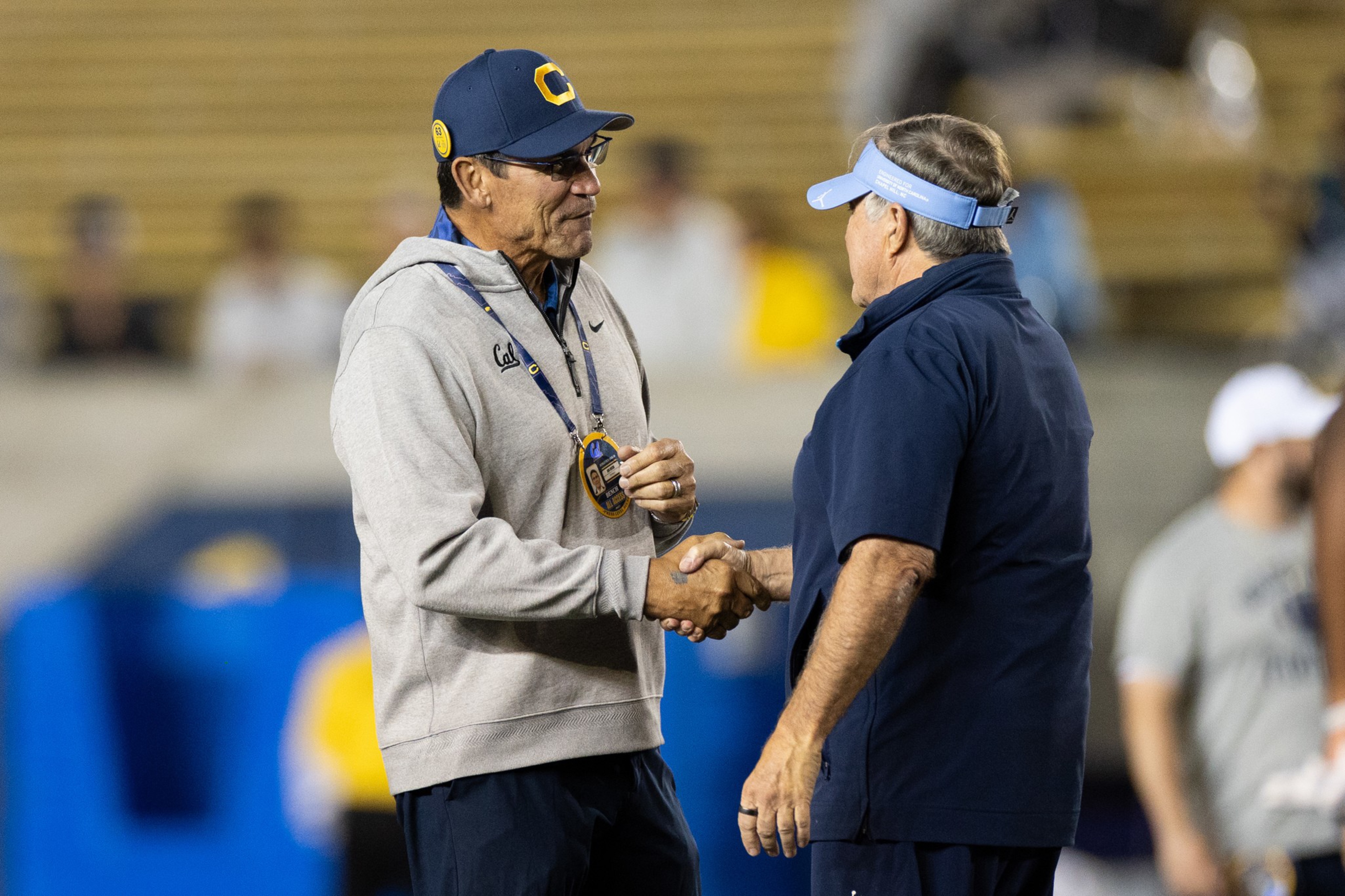 Two men in sportswear shake hands on a field, one wearing a gray hoodie and cap, the other in a dark blue shirt and light blue visor.