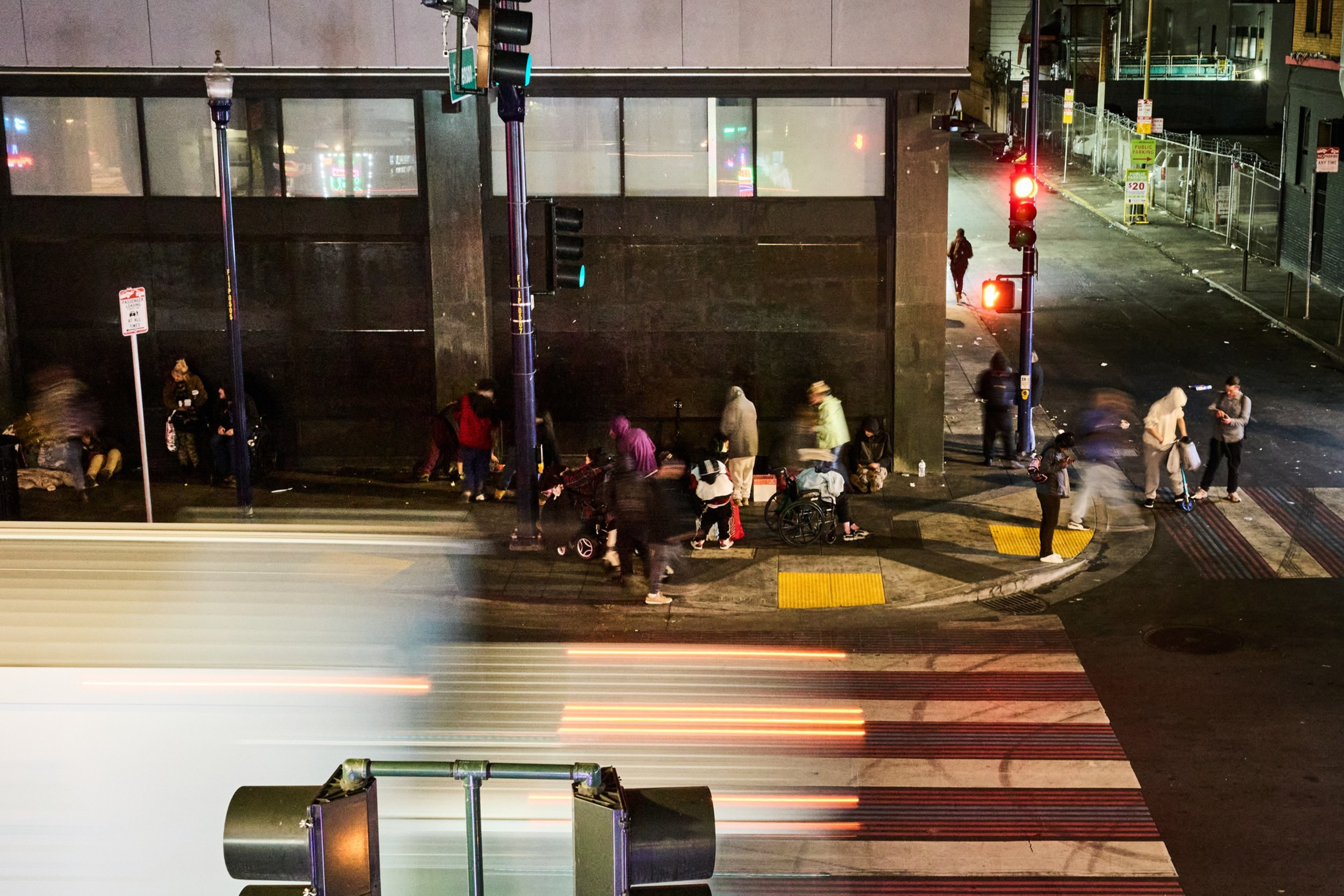 A busy street corner shows people standing and sitting by a building, some with strollers or wheelchairs. A streak of motion from a vehicle is visible in the foreground.