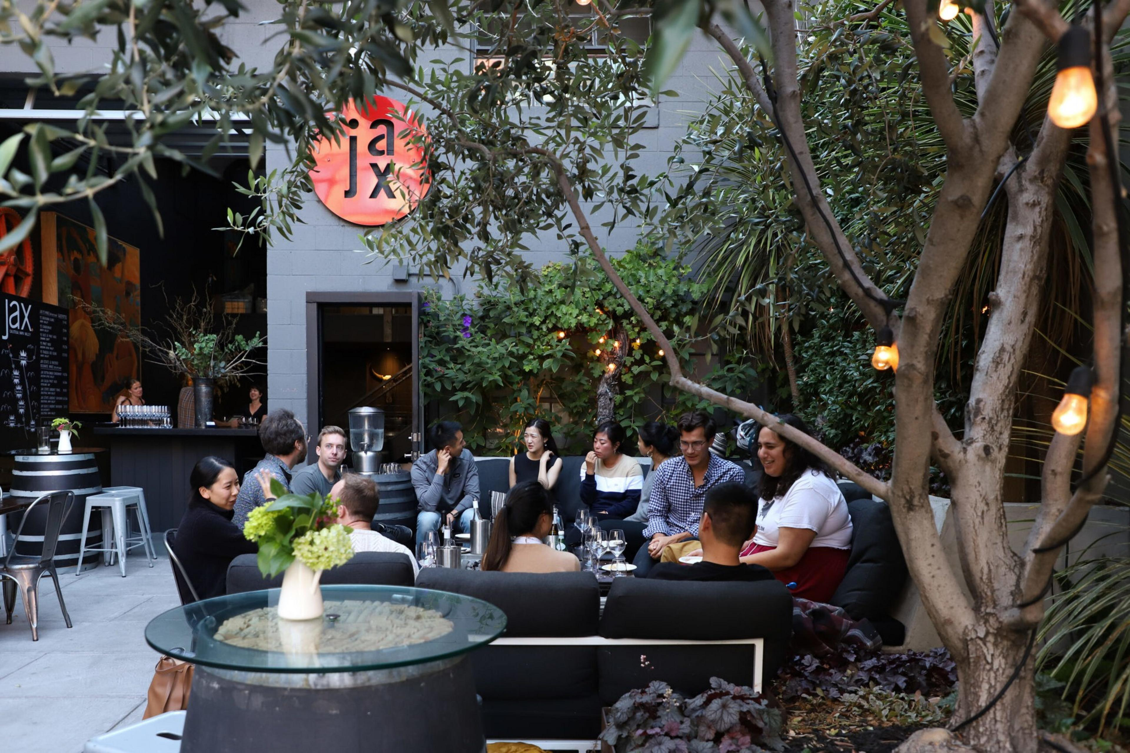 A group of people sits and chats in a cozy outdoor patio with greenery, string lights, and a bar area labeled "jax" in the background.
