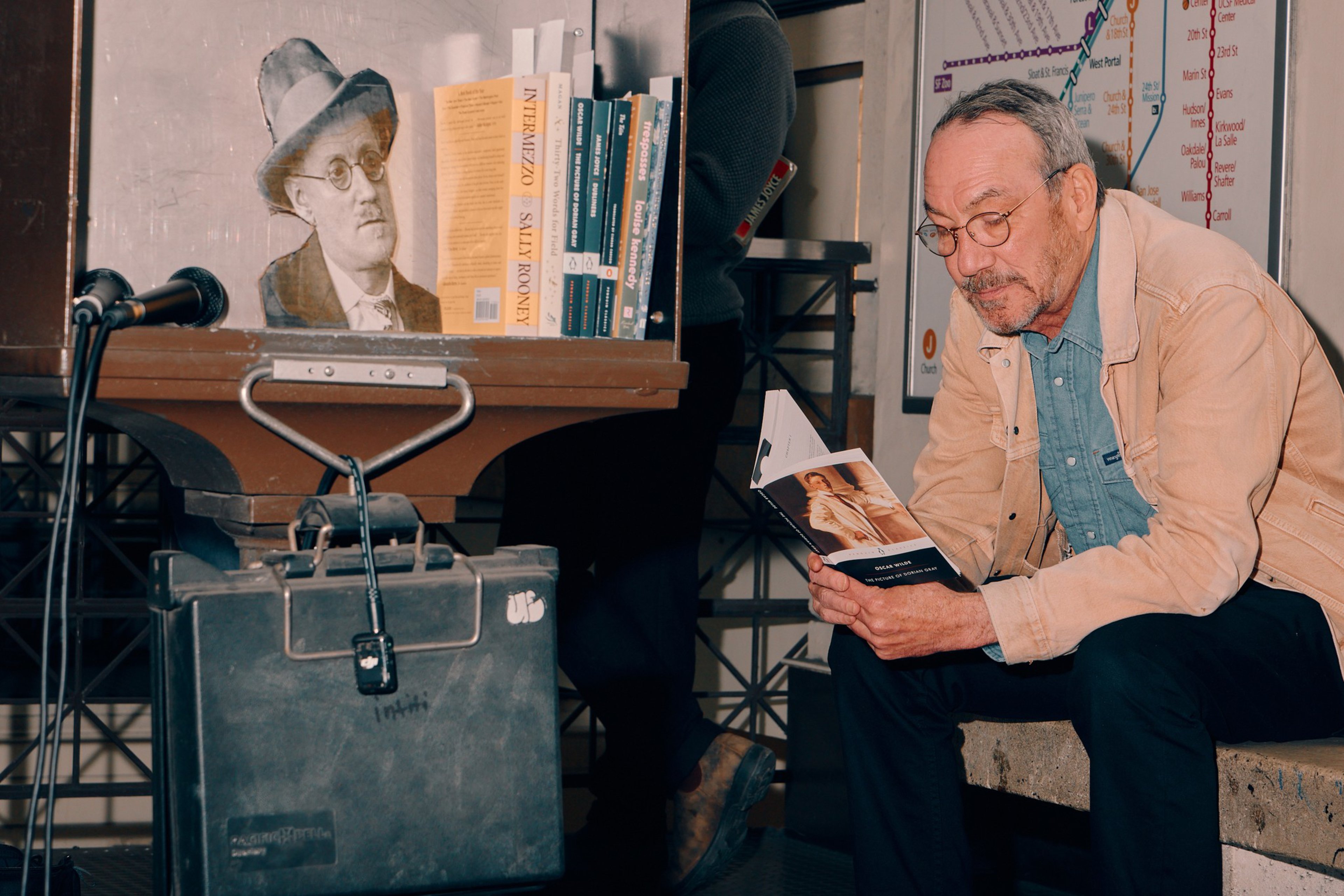 A man with glasses and a beige jacket sits reading a book near a shelf holding books and a black-and-white illustration of a man wearing a hat and round glasses.