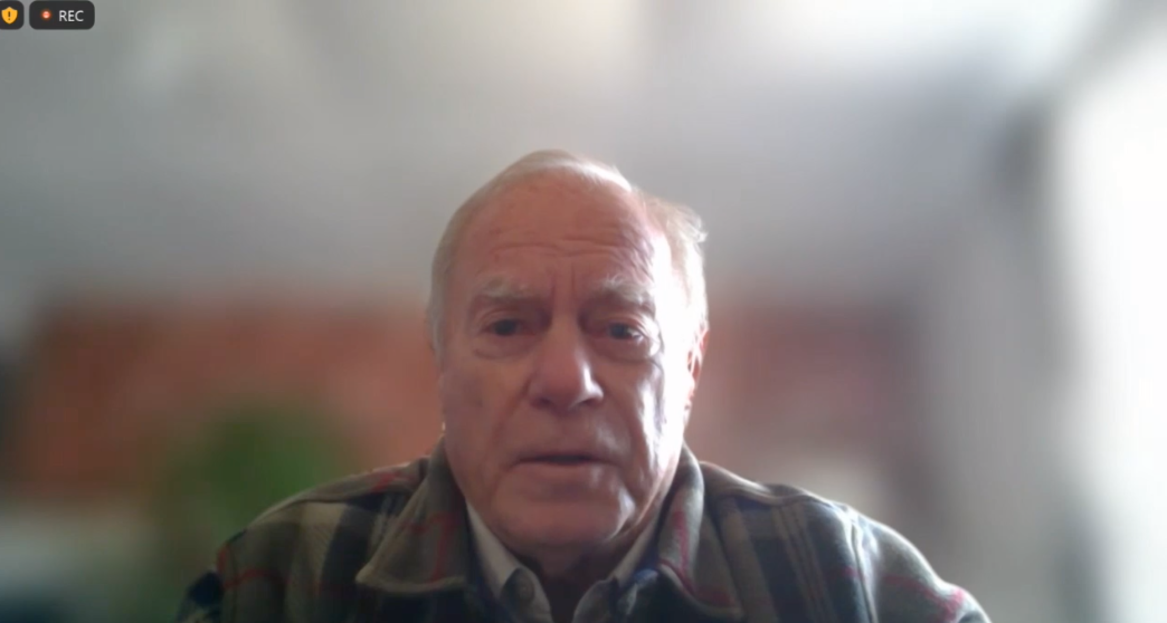 An older man with short white hair wears a plaid jacket and looks directly at the camera in a softly lit room with a blurred background.
