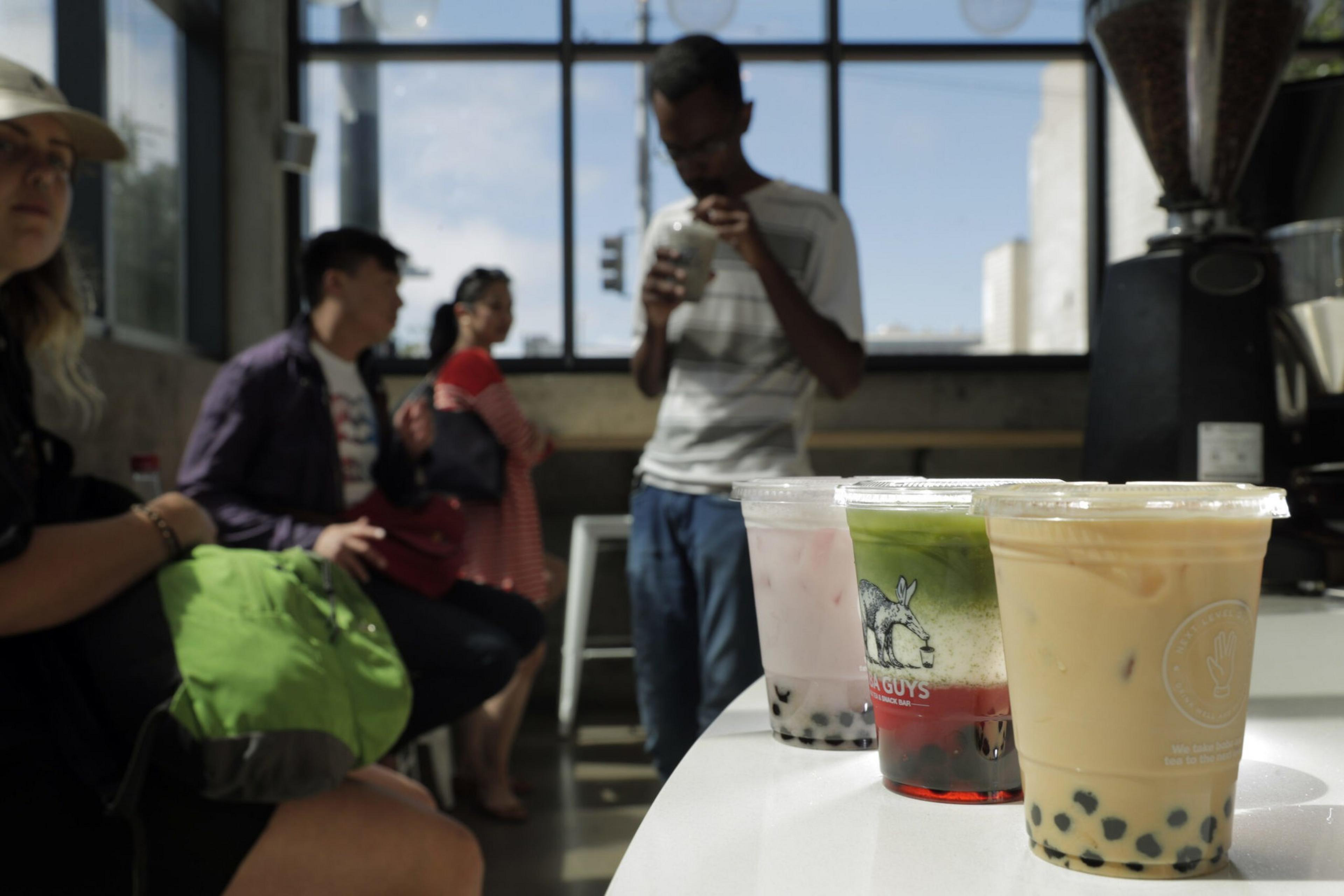 Three bubble tea drinks sit on a table, with people sitting and standing around in a bright cafÃ©. A large window in the background lets in natural light.