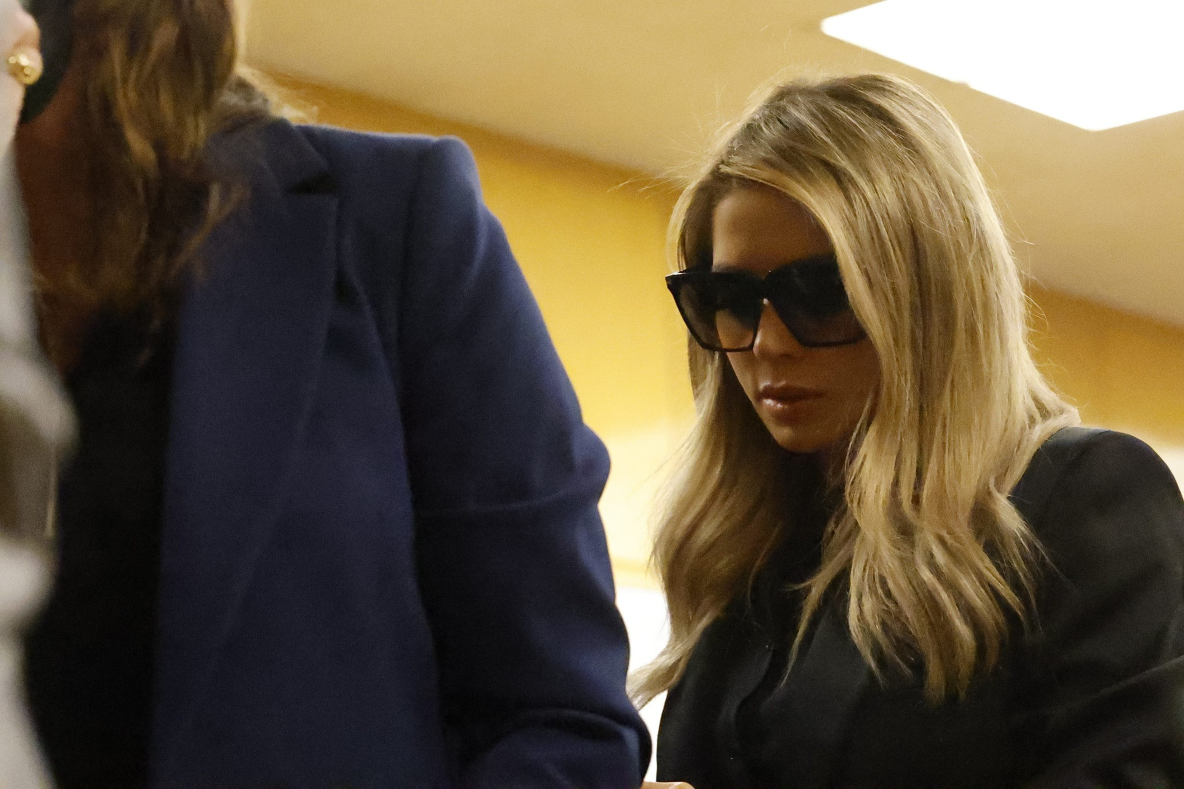 A woman with long blonde hair wears large dark sunglasses and a black outfit, standing next to another person in a blue jacket. The background is softly lit.