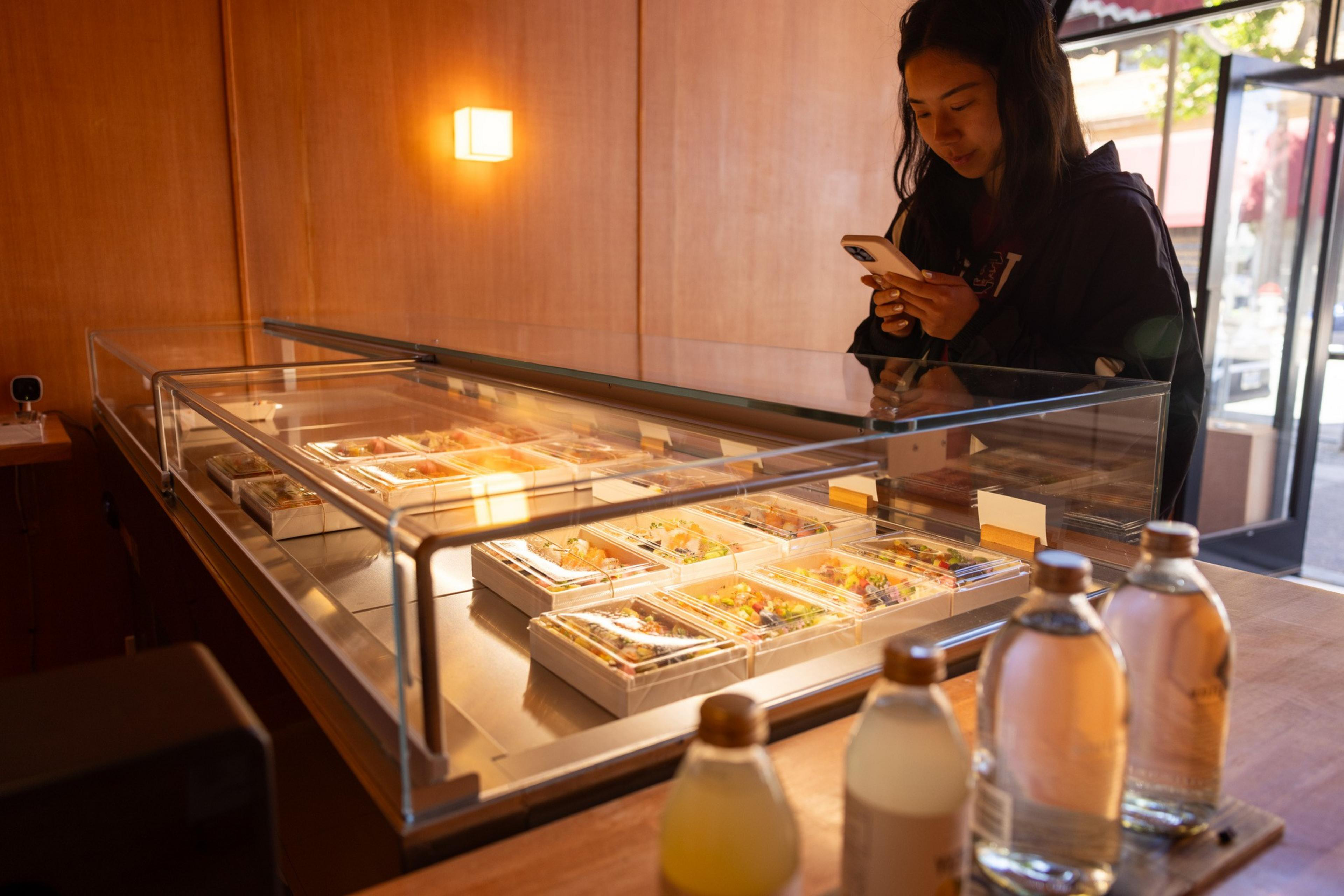 A woman stands in front of a glass display case filled with neatly arranged food containers, holding her phone. Bottles of beverages are on the counter nearby.