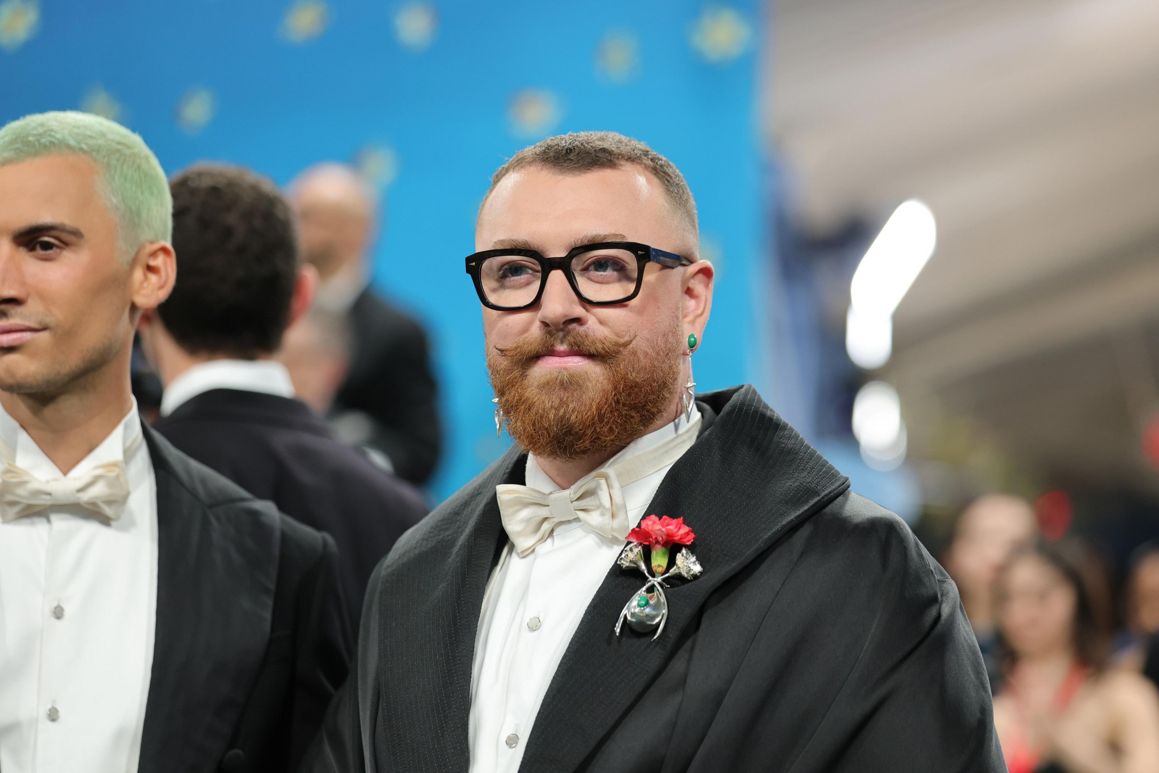 A bearded man with glasses, a white bow tie, and a dark suit jacket adorned with a red flower pin stands next to another man with mint green hair.