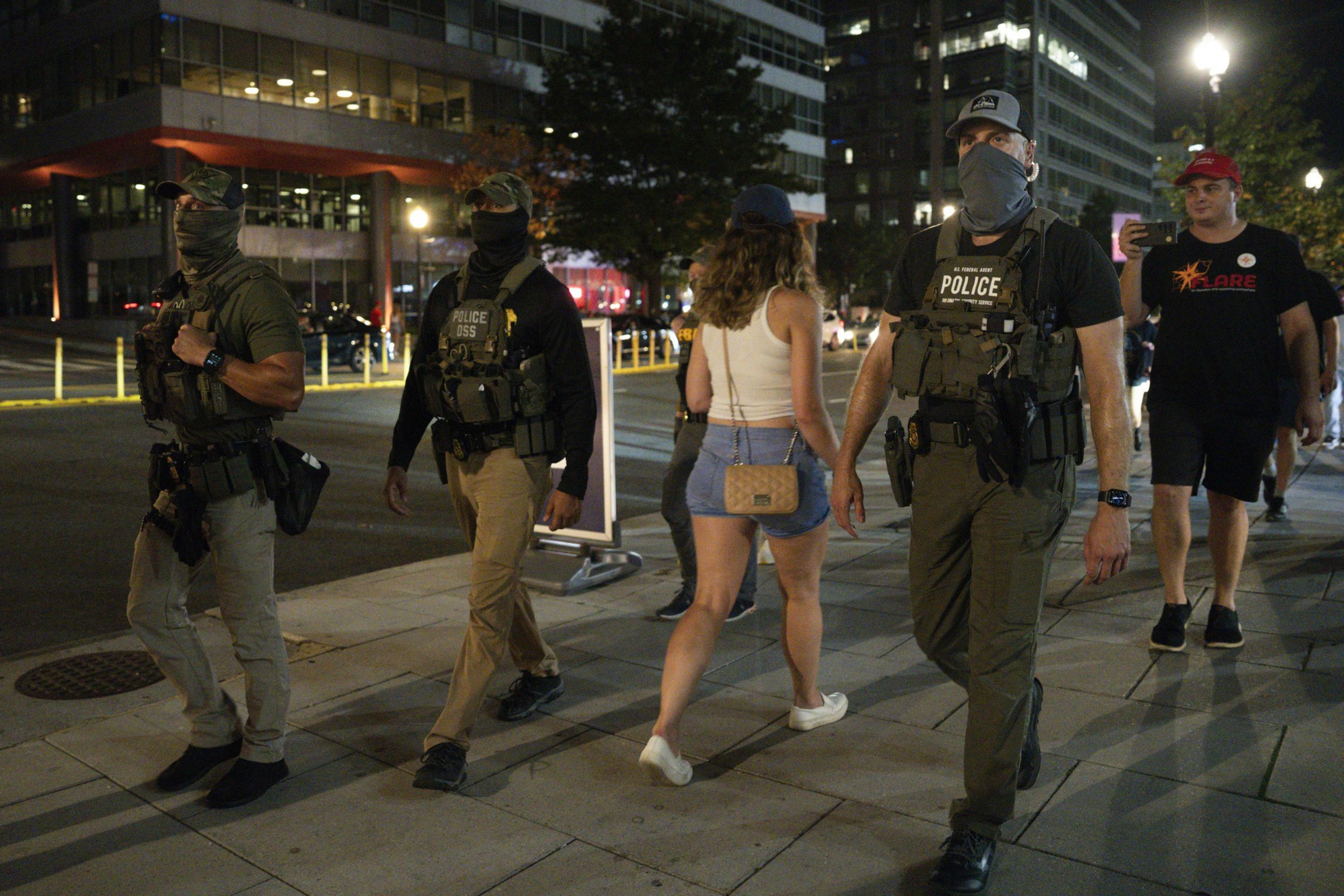 Three armed police officers wearing tactical gear and face coverings walk on a city sidewalk at night alongside casually dressed pedestrians.