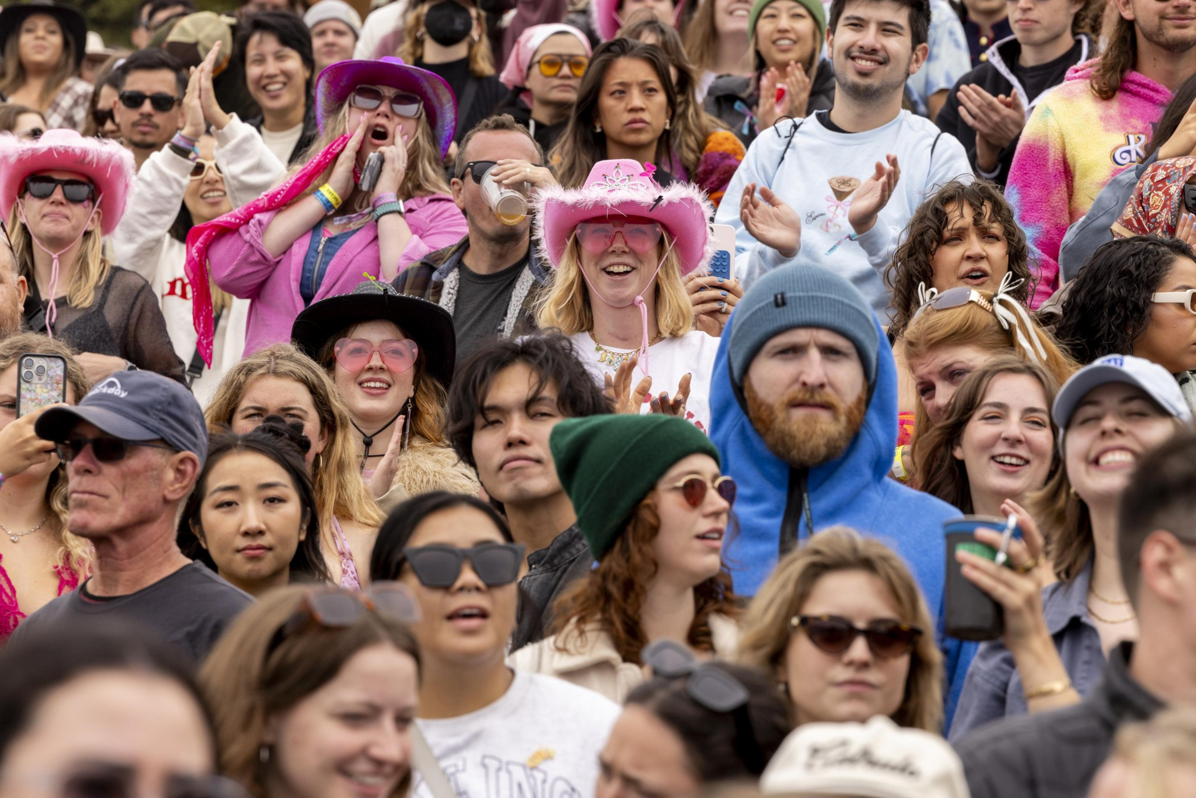 A lively crowd of people, many wearing colorful hats and sunglasses, are gathered outdoors, smiling and clapping, enjoying a festive event.