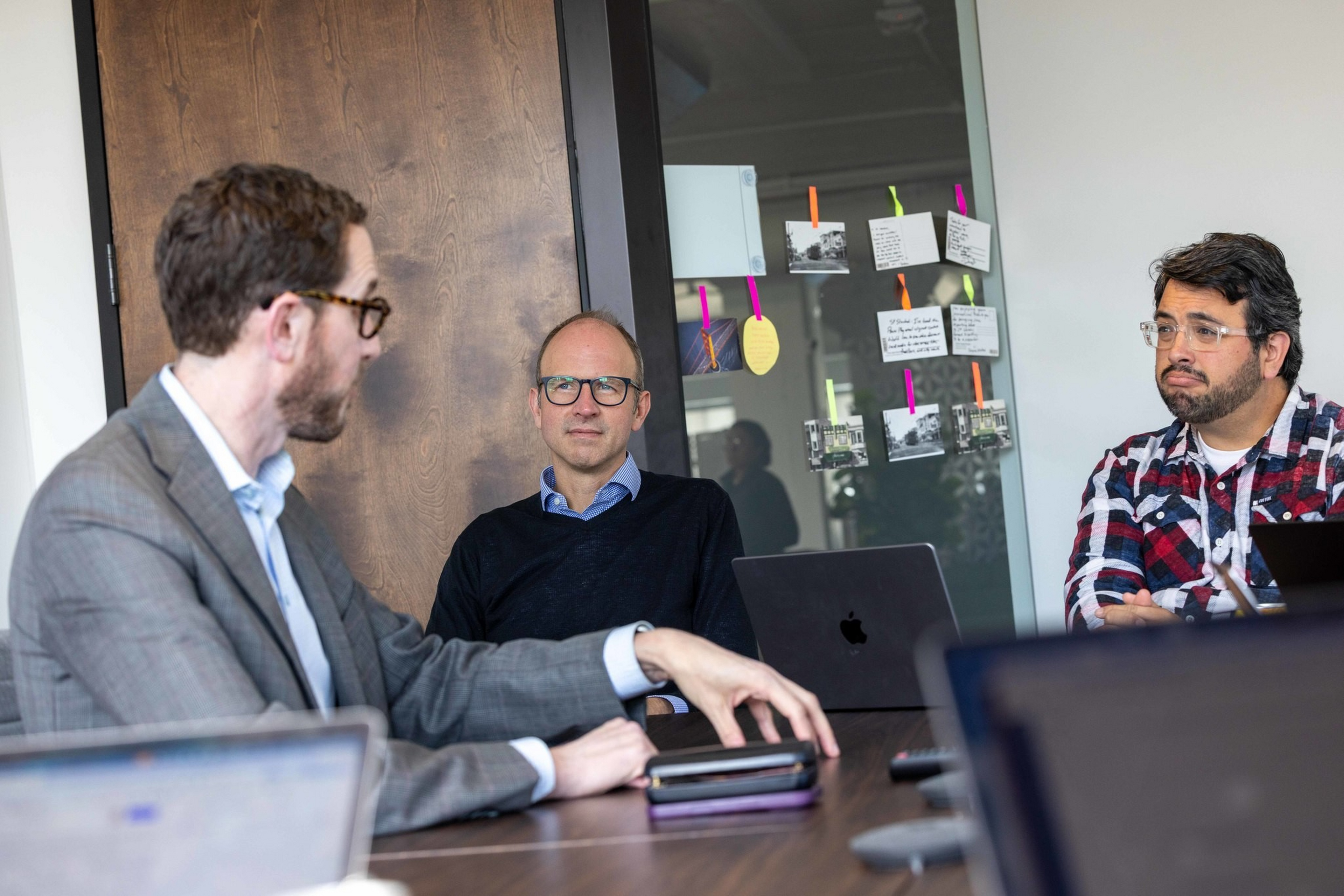 Three men sit around a conference table, two with laptops open, one in a plaid shirt, one in a suit, and one in a dark sweater with glasses.