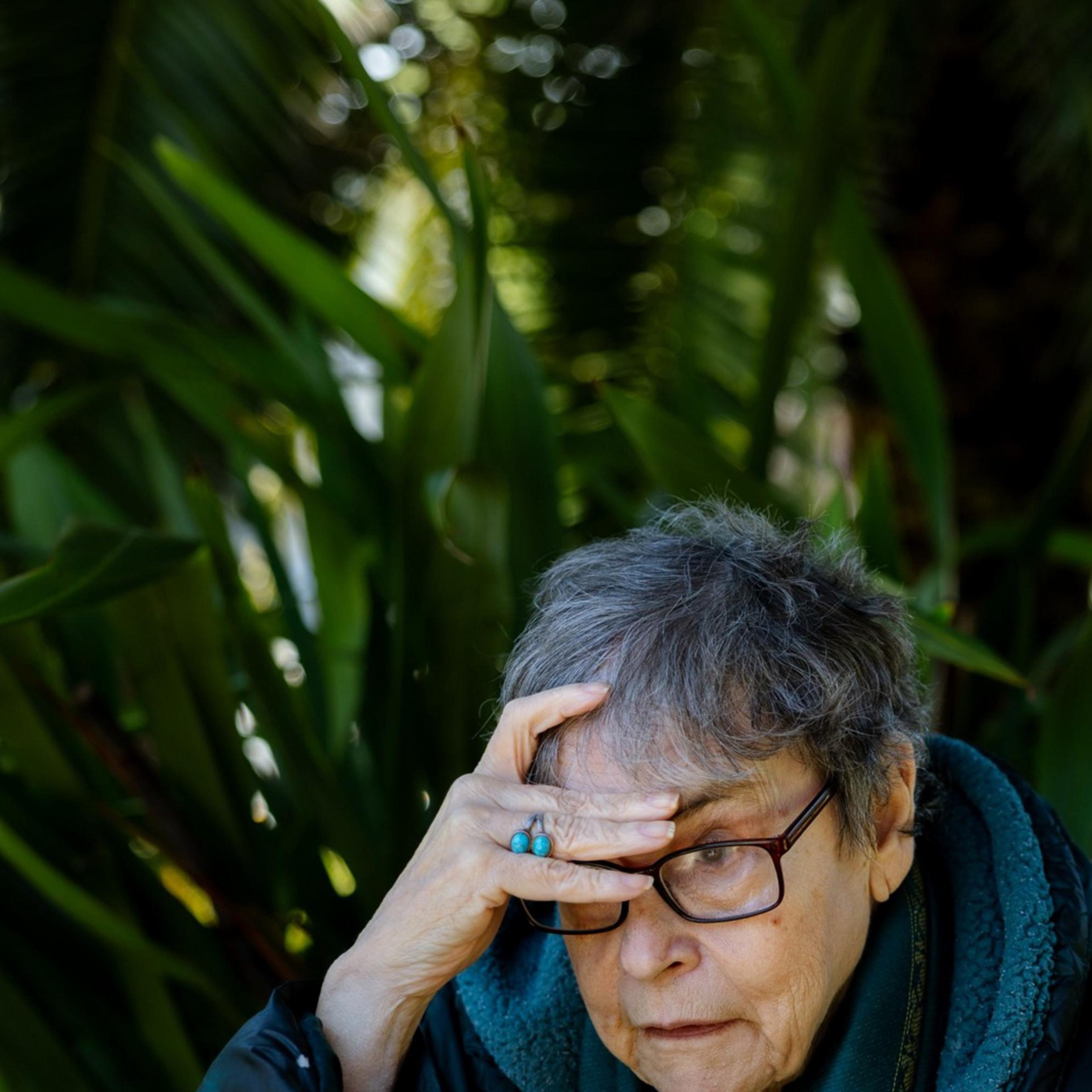 An older person with glasses looks thoughtful while resting their hand on their forehead. They sit outdoors surrounded by large green leaves.