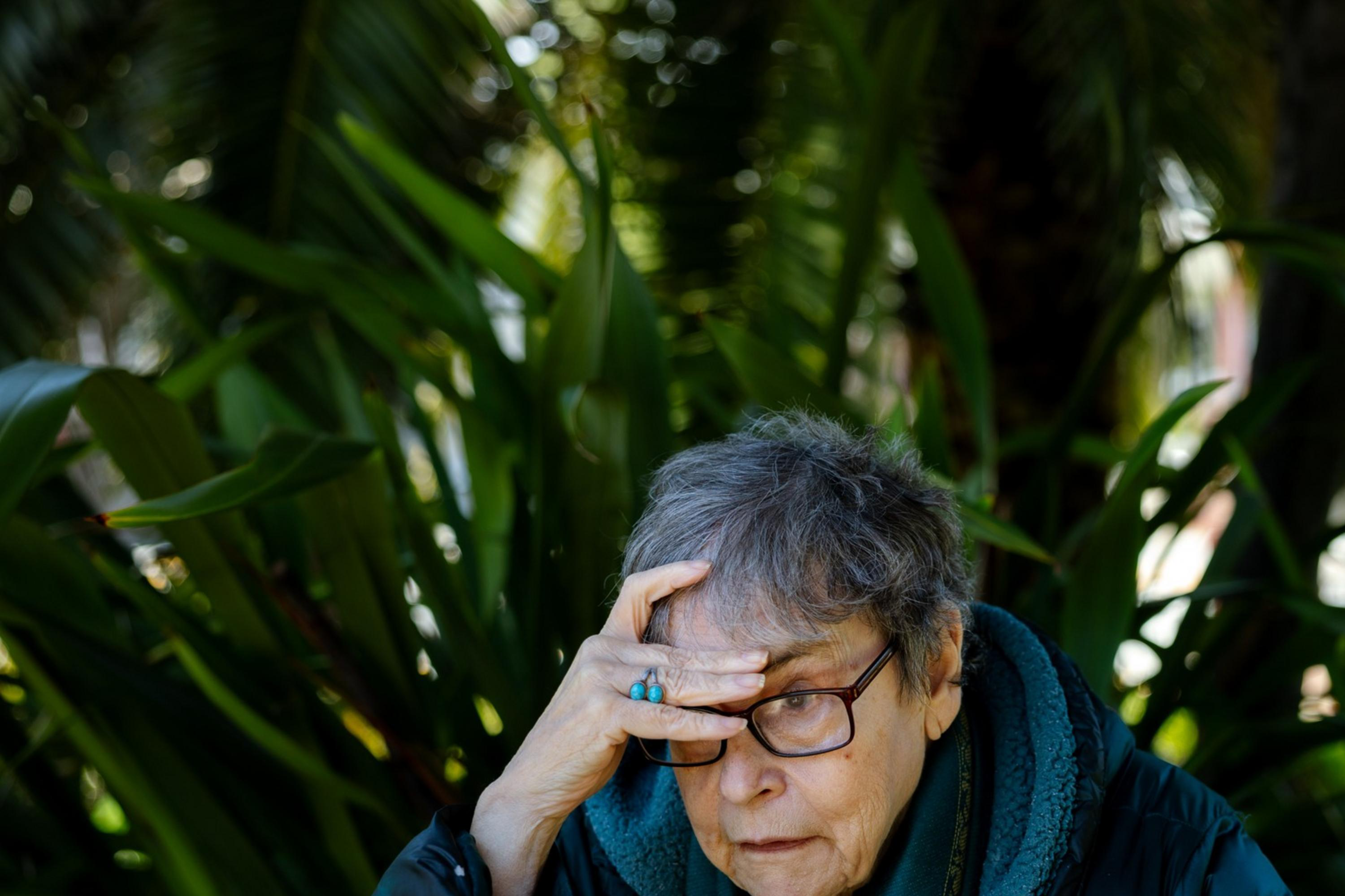 An older person with glasses looks thoughtful while resting their hand on their forehead. They sit outdoors surrounded by large green leaves.