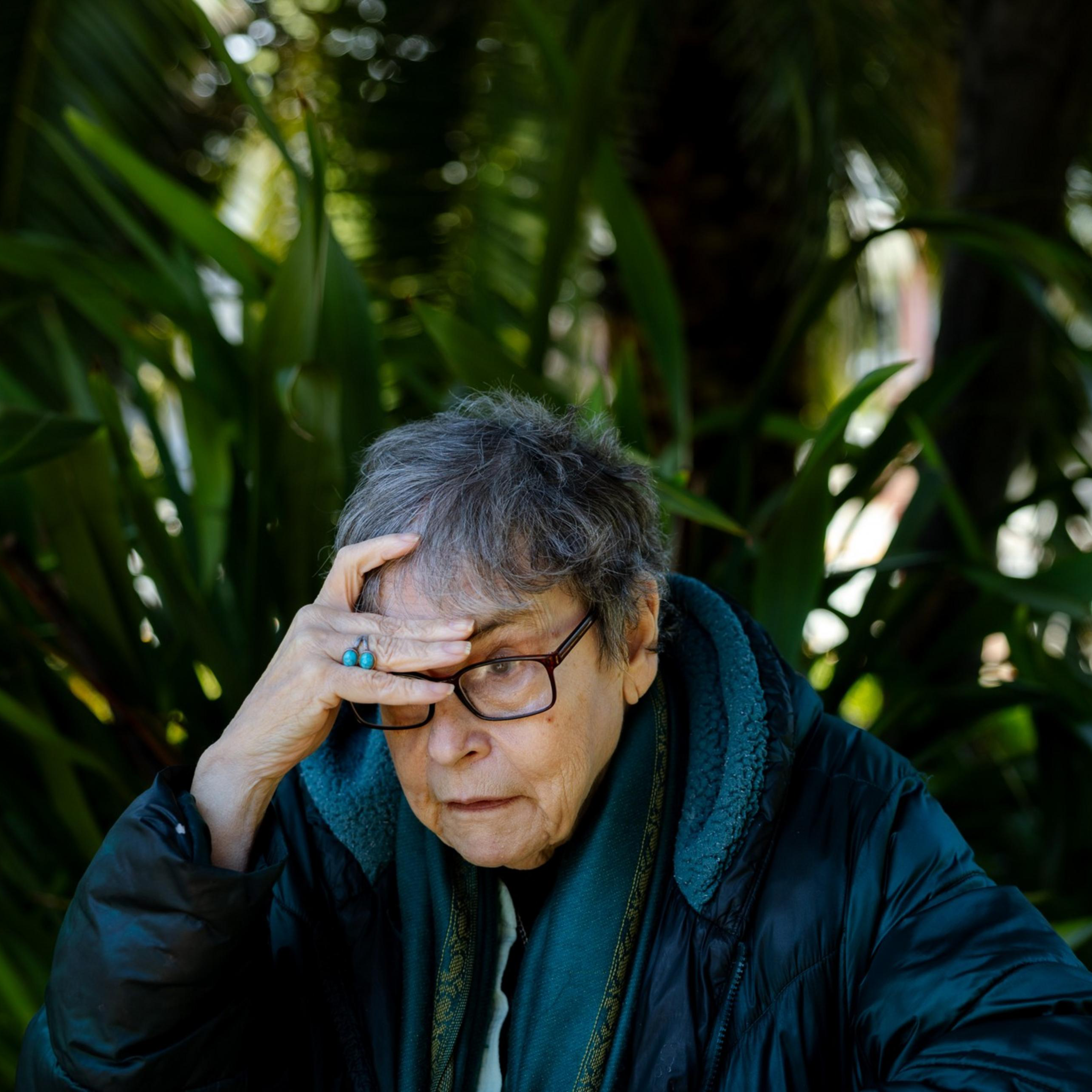 An older person with glasses looks thoughtful while resting their hand on their forehead. They sit outdoors surrounded by large green leaves.