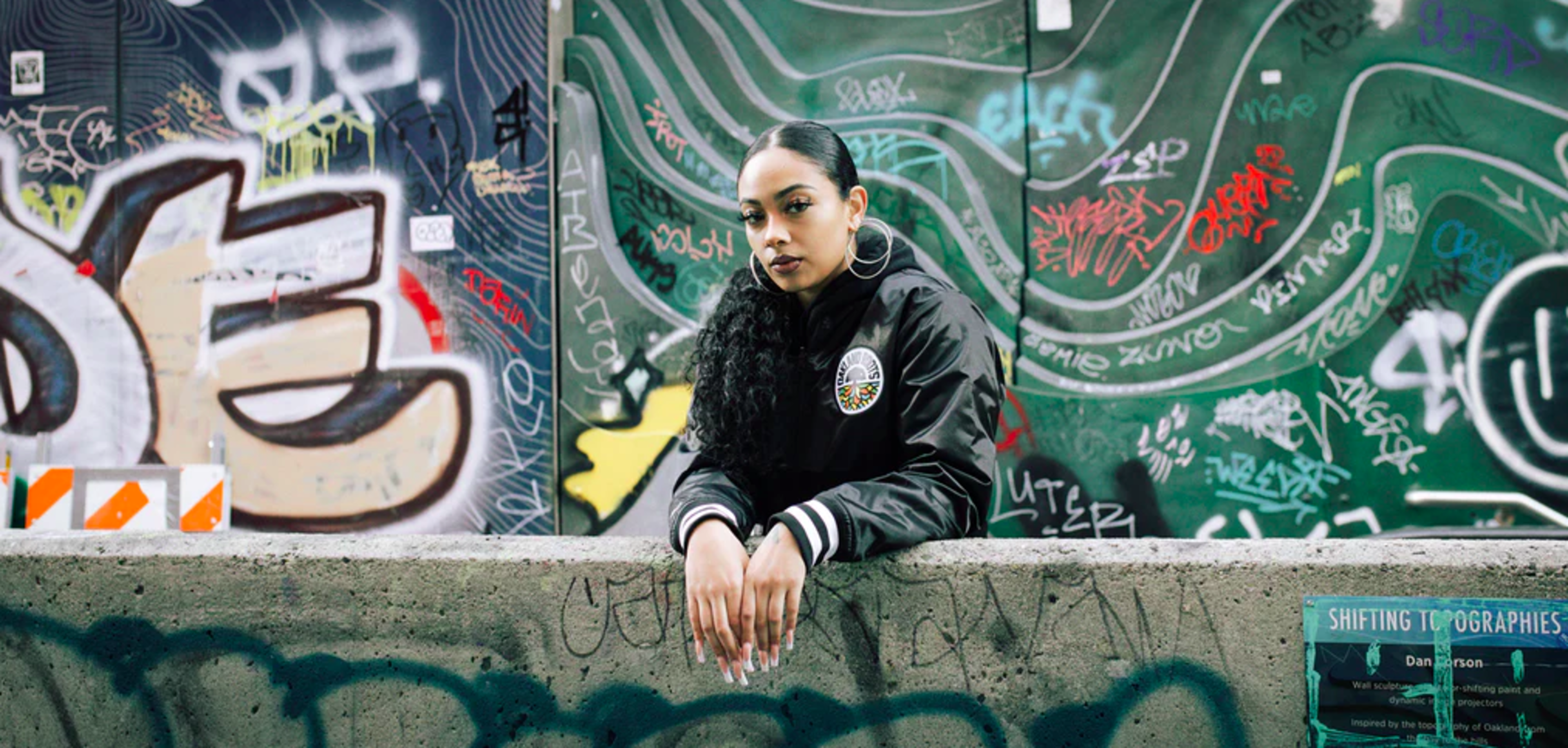 A woman in a black jacket leans on a graffiti-covered concrete barrier, with colorful graffiti art on a wall behind her.