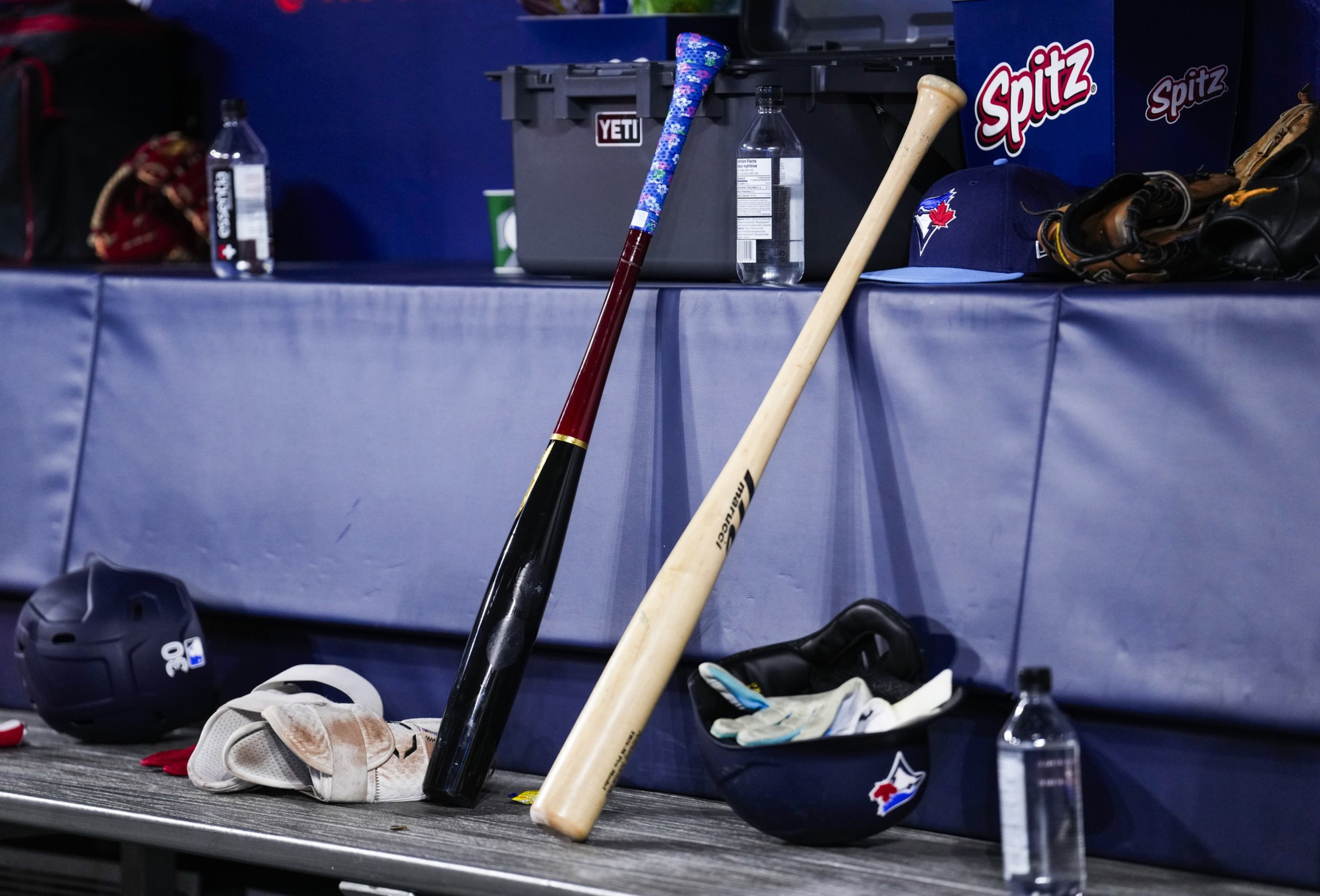 Two baseball bats leaning against a padded bench with gloves, helmets, water bottles, and a cap nearby, set in a dugout with branded boxes.