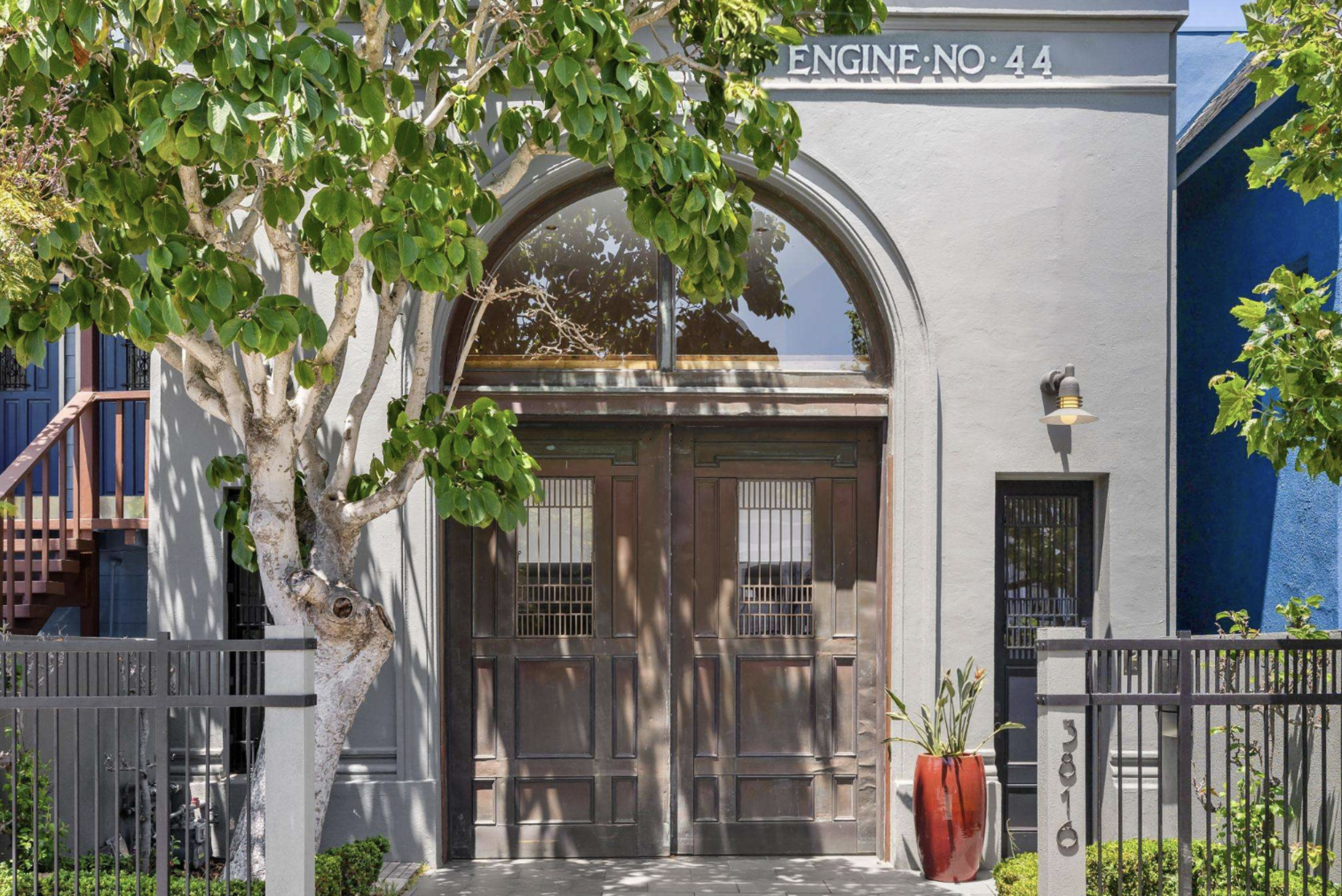 A gray building with large wooden double doors beneath an arched window, labeled “ENGINE NO. 44,” flanked by a tree and a red planter.