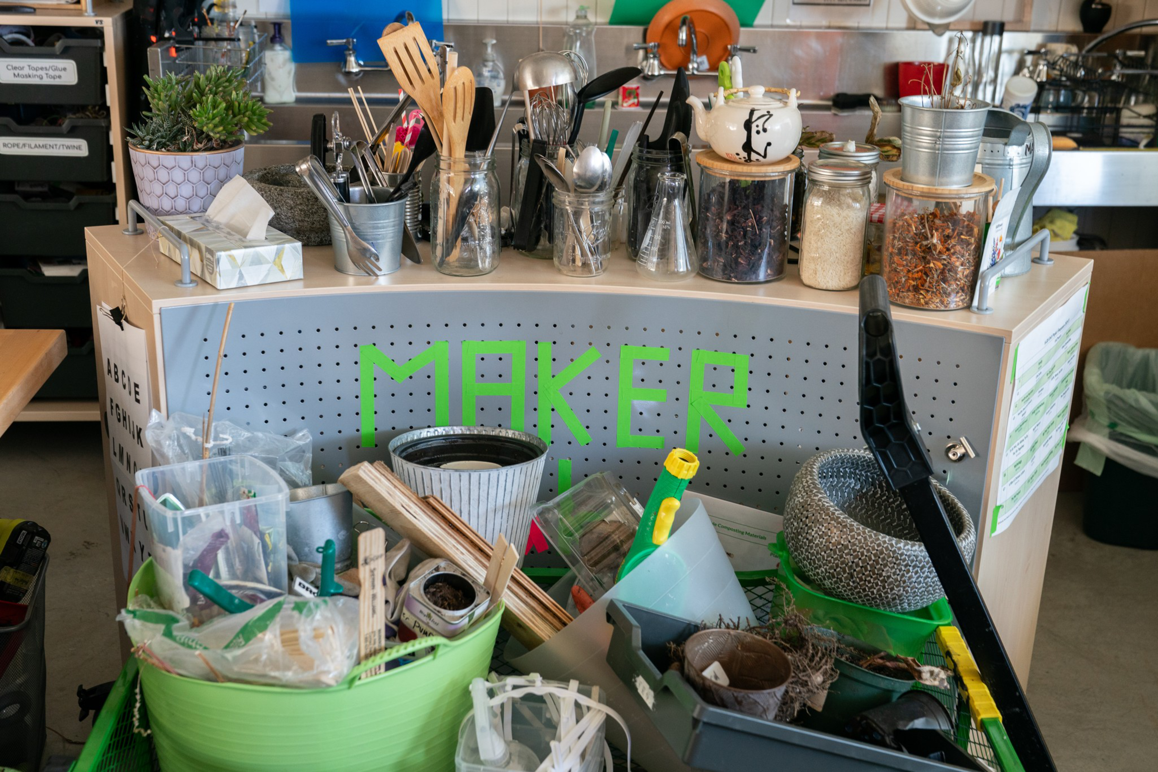 The image shows a cluttered workspace labeled &quot;MAKER,&quot; with various utensils, jars, plants, and crafting supplies organized on a table and in bins.