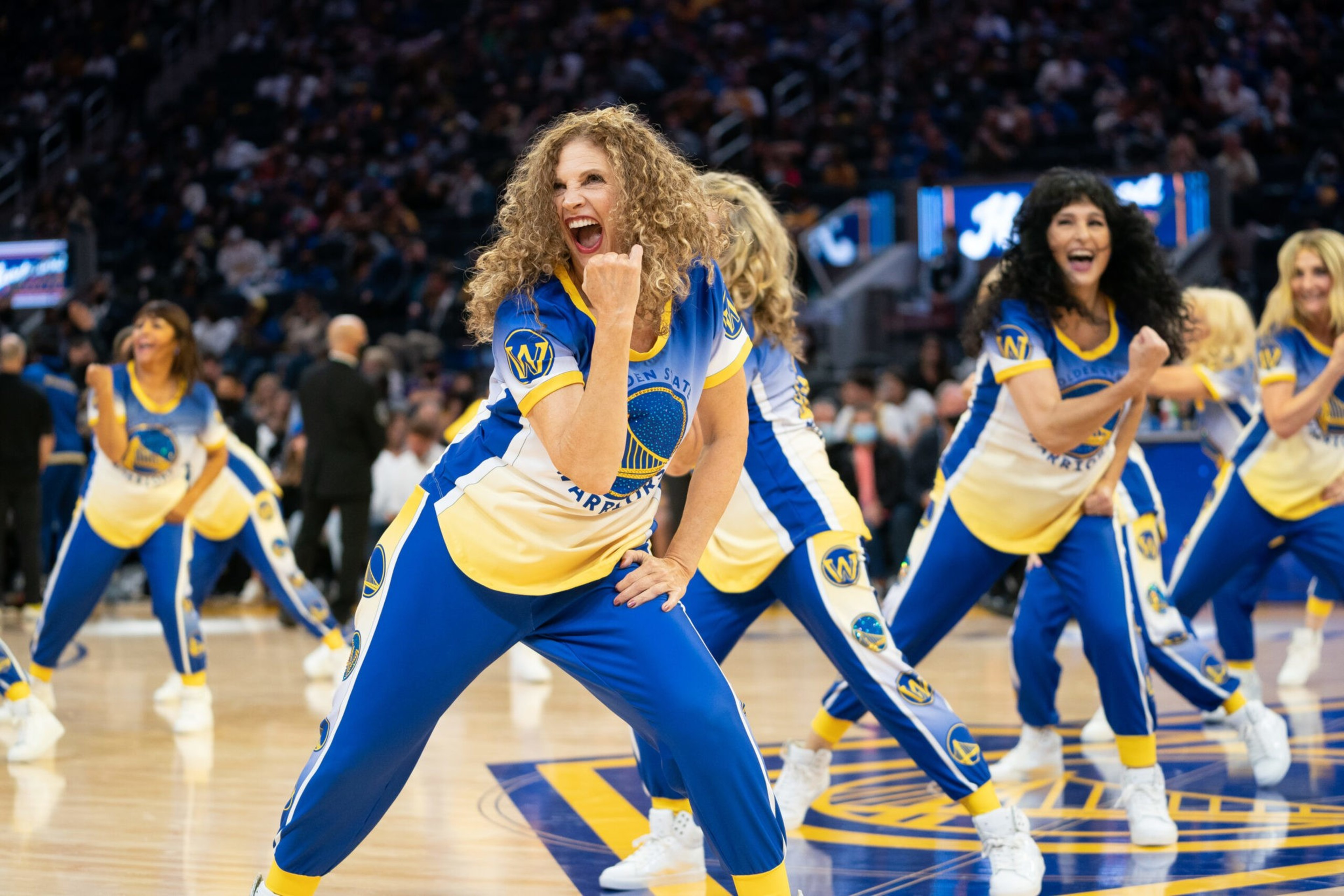 Women in blue and yellow Golden State Warriors outfits are energetically dancing on a basketball court during a game.