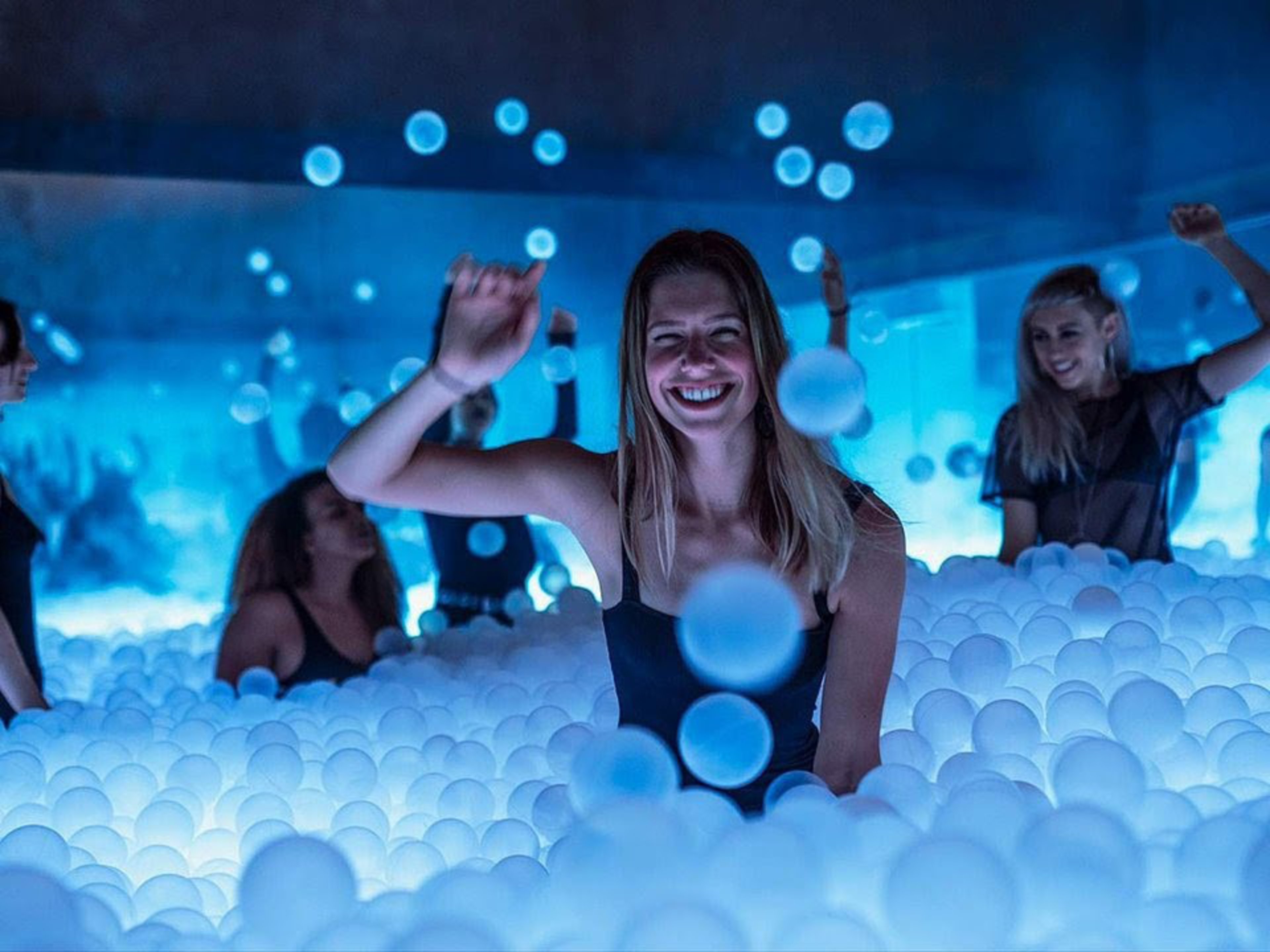 A group of people are playing and smiling in a glowing blue ball pit filled with white balls in a dimly lit room.