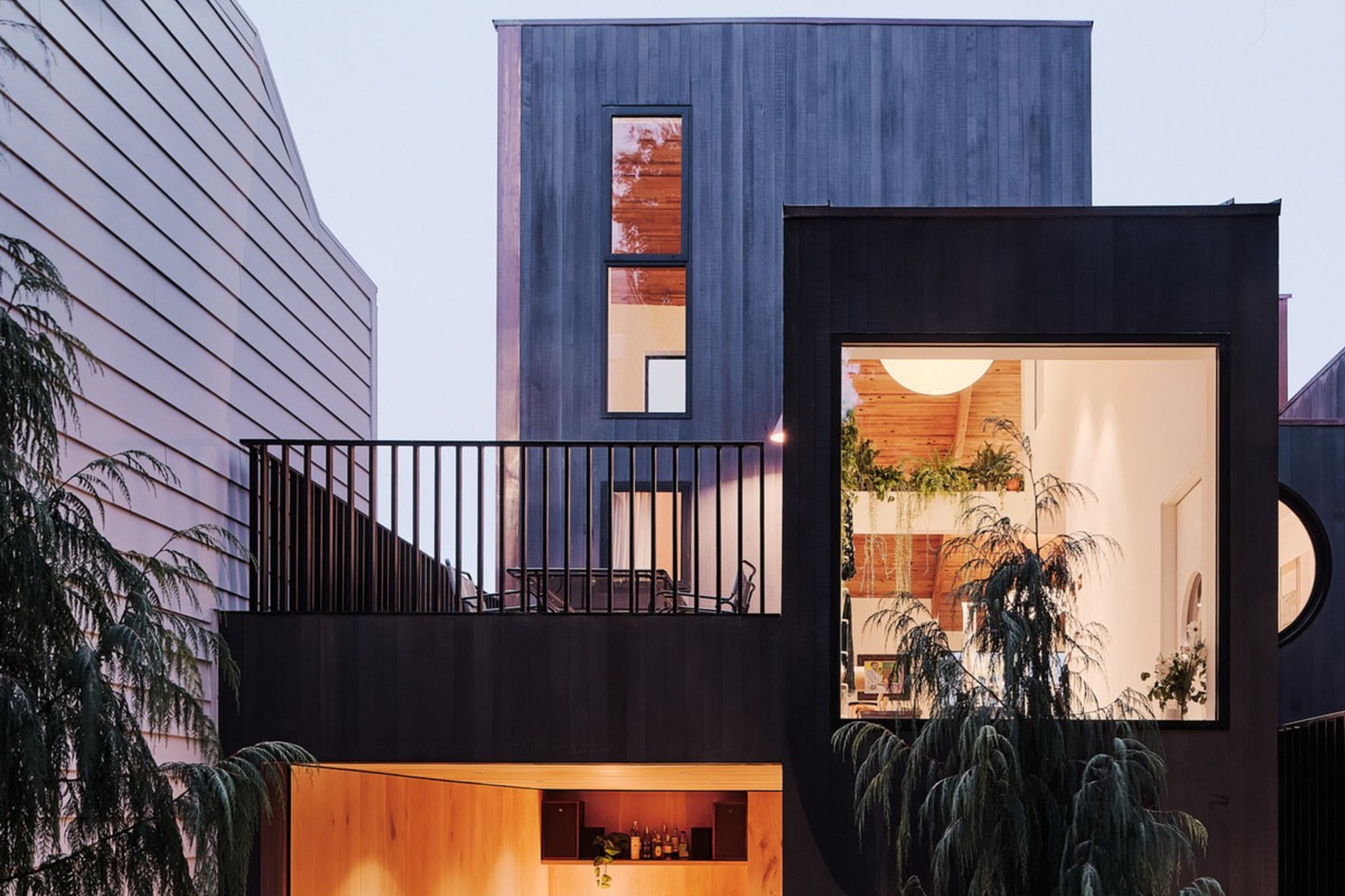 A modern, two-story house with large windows is surrounded by trees. The interior shows a cozy living area and a warm, softly lit kitchen.