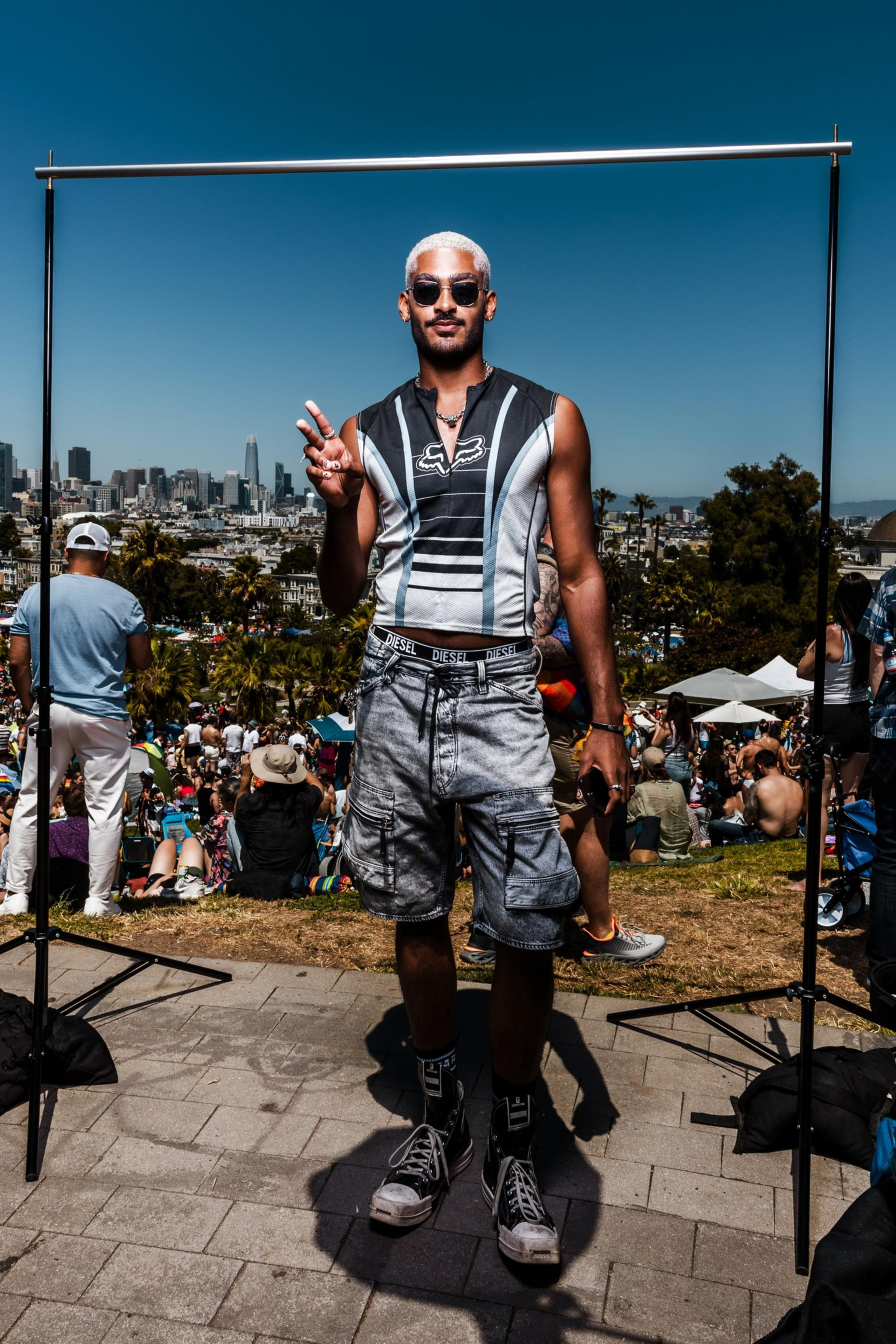 A person stands outdoors in front of a crowd, making a peace sign. They wear sunglasses, a sleeveless top, cargo shorts, and high-top sneakers, with a city skyline behind them.