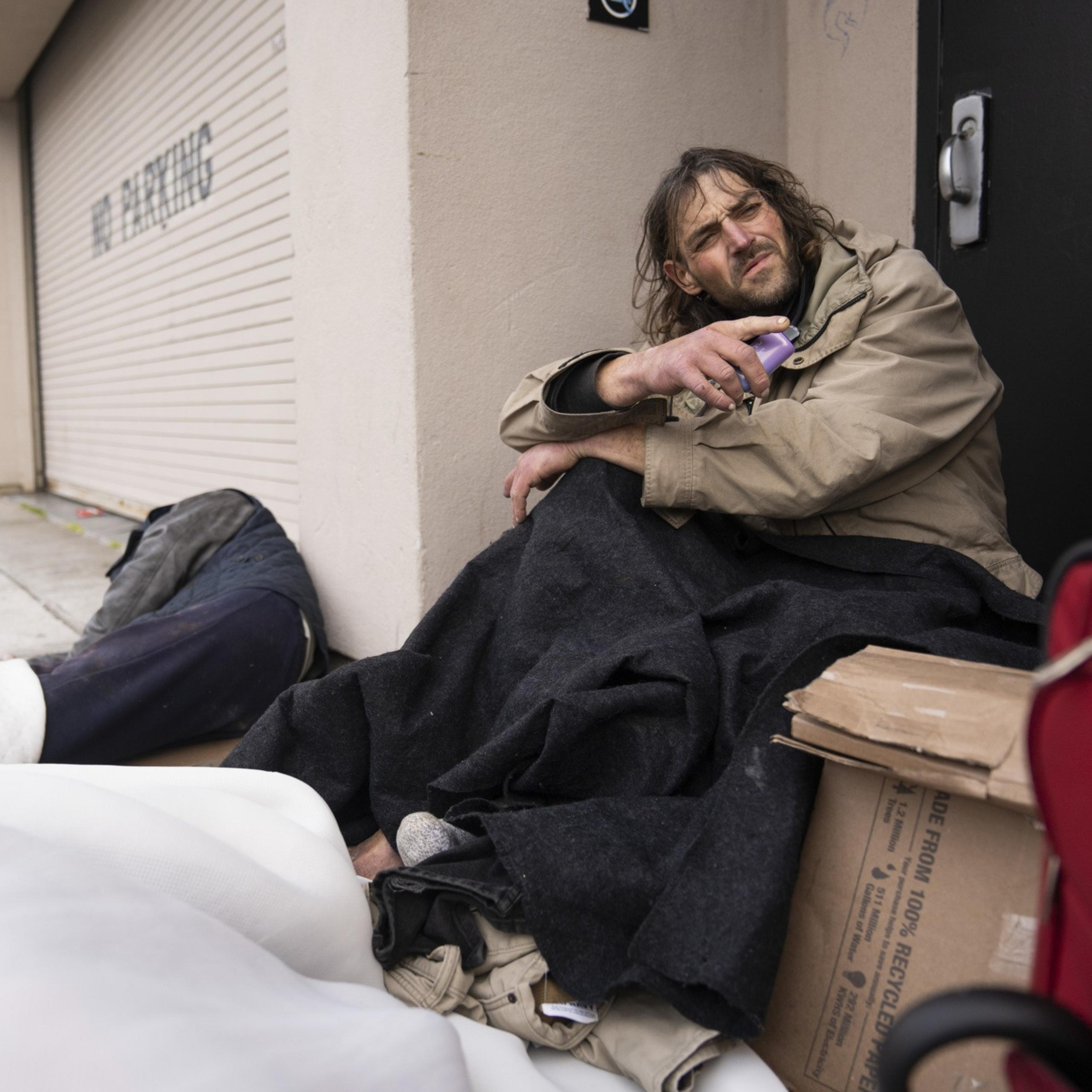 A man sits on the sidewalk wrapped in a blanket, holding an object, beside a closed garage door. Nearby, there's a red suitcase and cardboard boxes.
