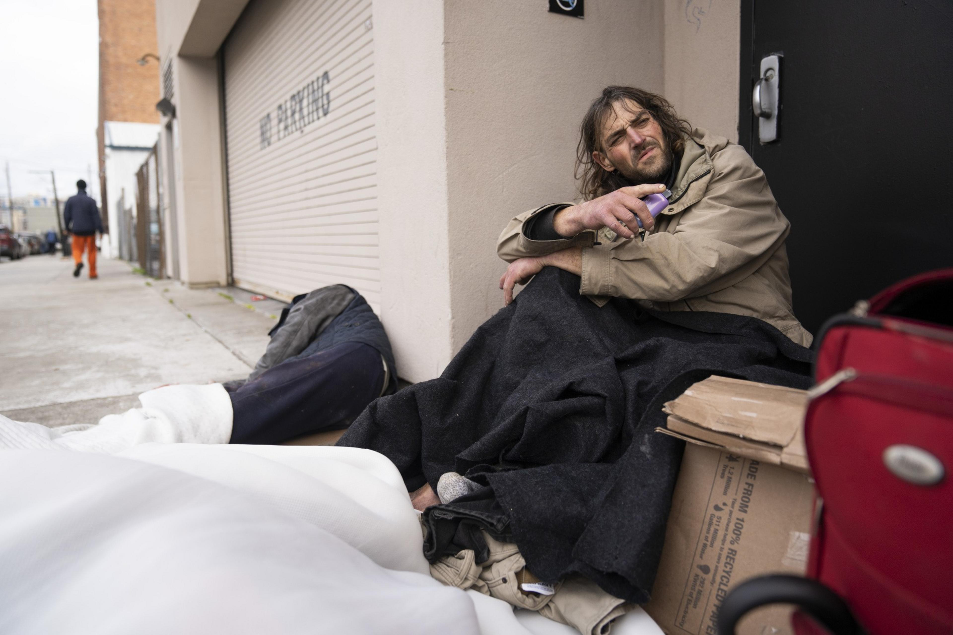 A man sits on the sidewalk wrapped in a blanket, holding an object, beside a closed garage door. Nearby, there's a red suitcase and cardboard boxes.