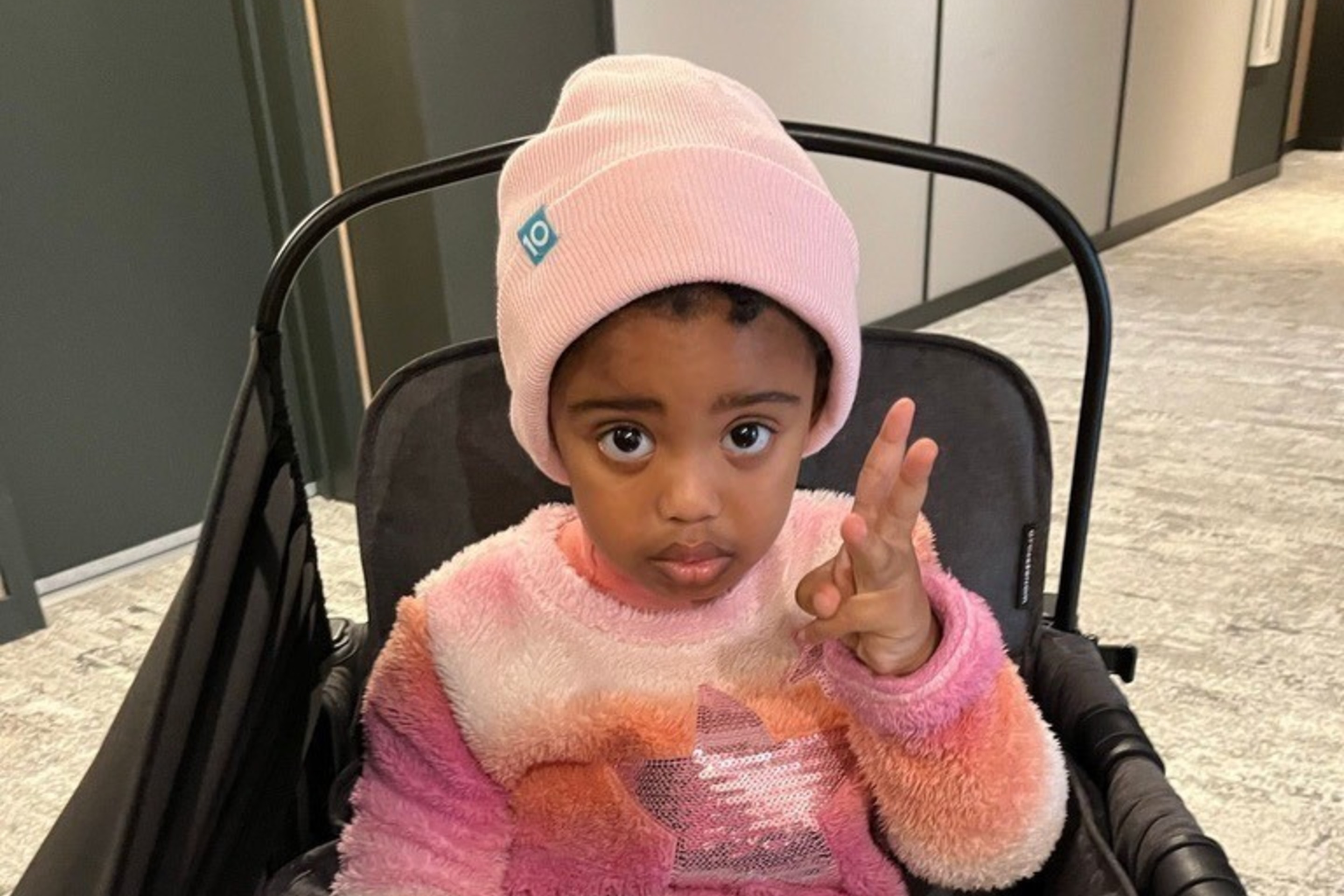 A young child wearing a pink beanie and a colorful sweater is sitting in a stroller, holding one hand up. An iPad with a kids' app is on their lap.