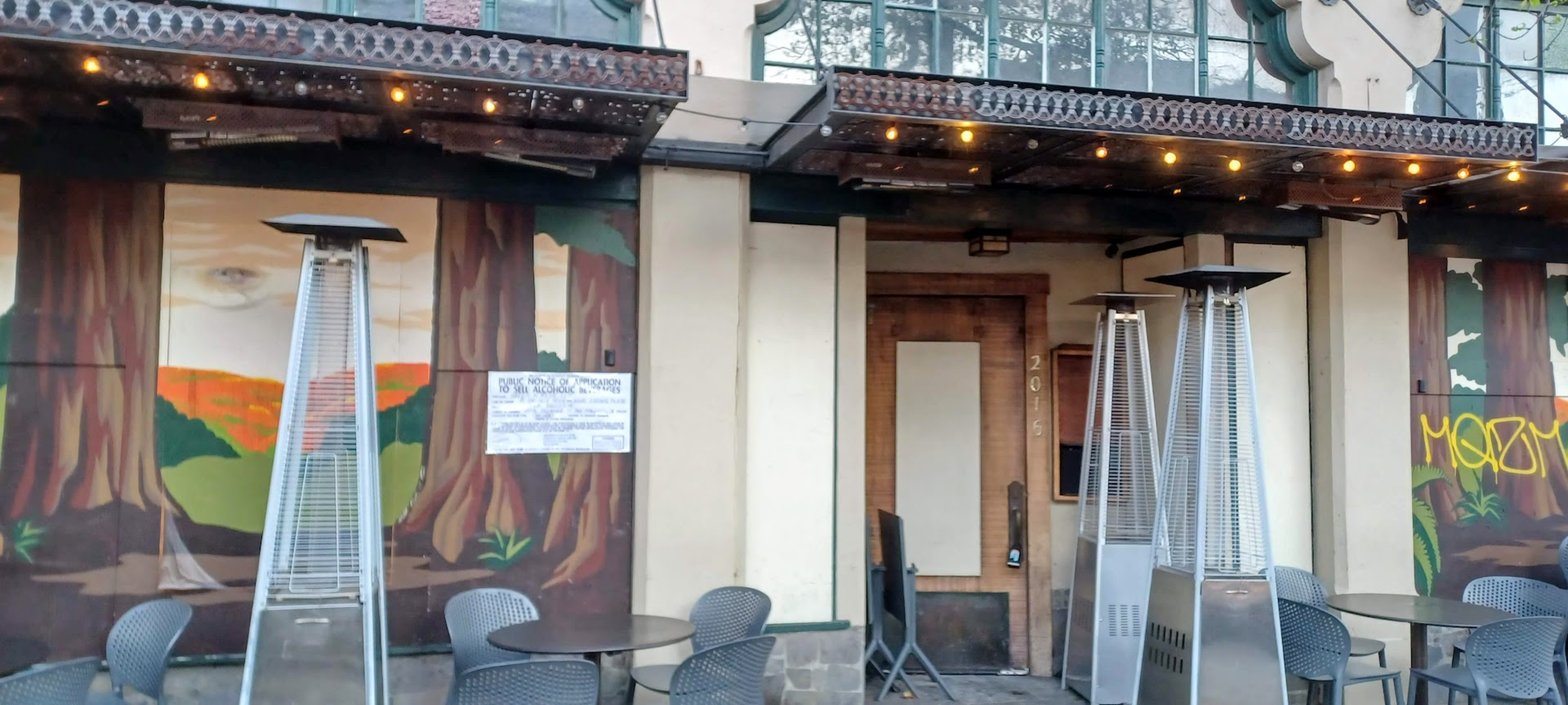 Empty outdoor seating with tables, chairs, and tall patio heaters stands in front of a closed wooden door and windows painted with forest murals, some graffiti visible.