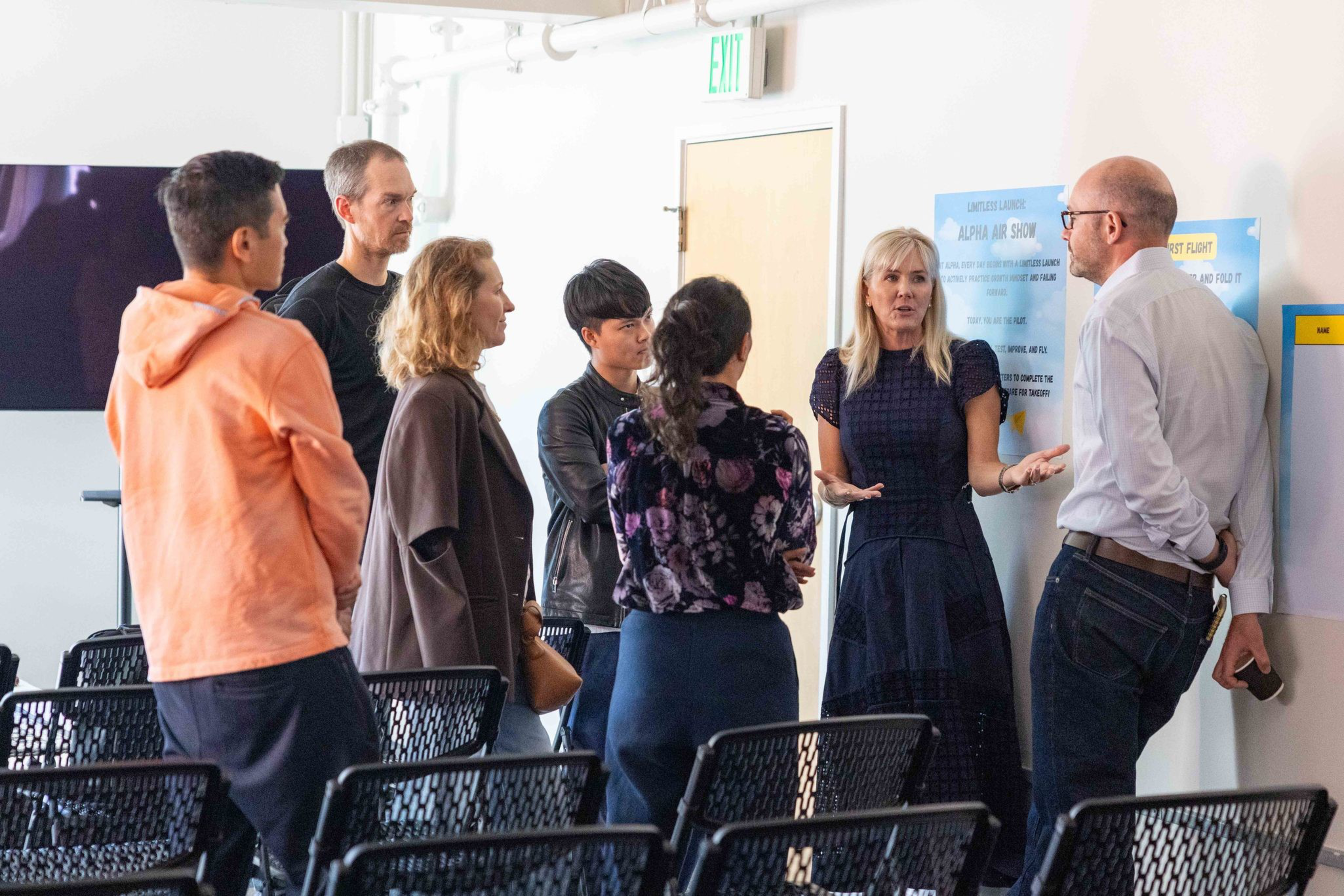 Six people stand and listen to a woman speaking near a wall with posters, while empty chairs face them in a bright room.