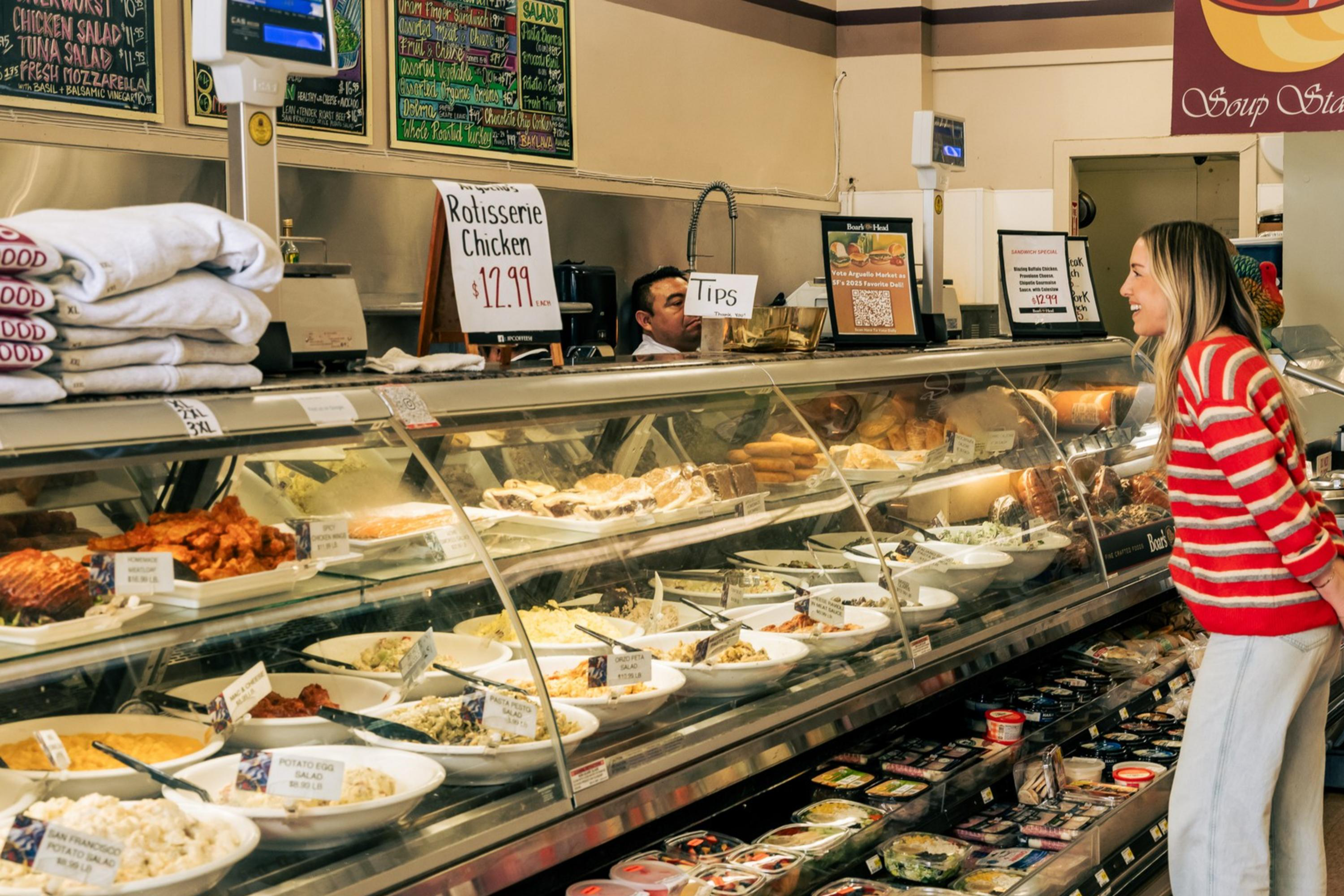 A woman in a red striped sweater examines a deli counter filled with various dishes. The sign above advertises rotisserie chicken for $12.99. The setting is a grocery store.