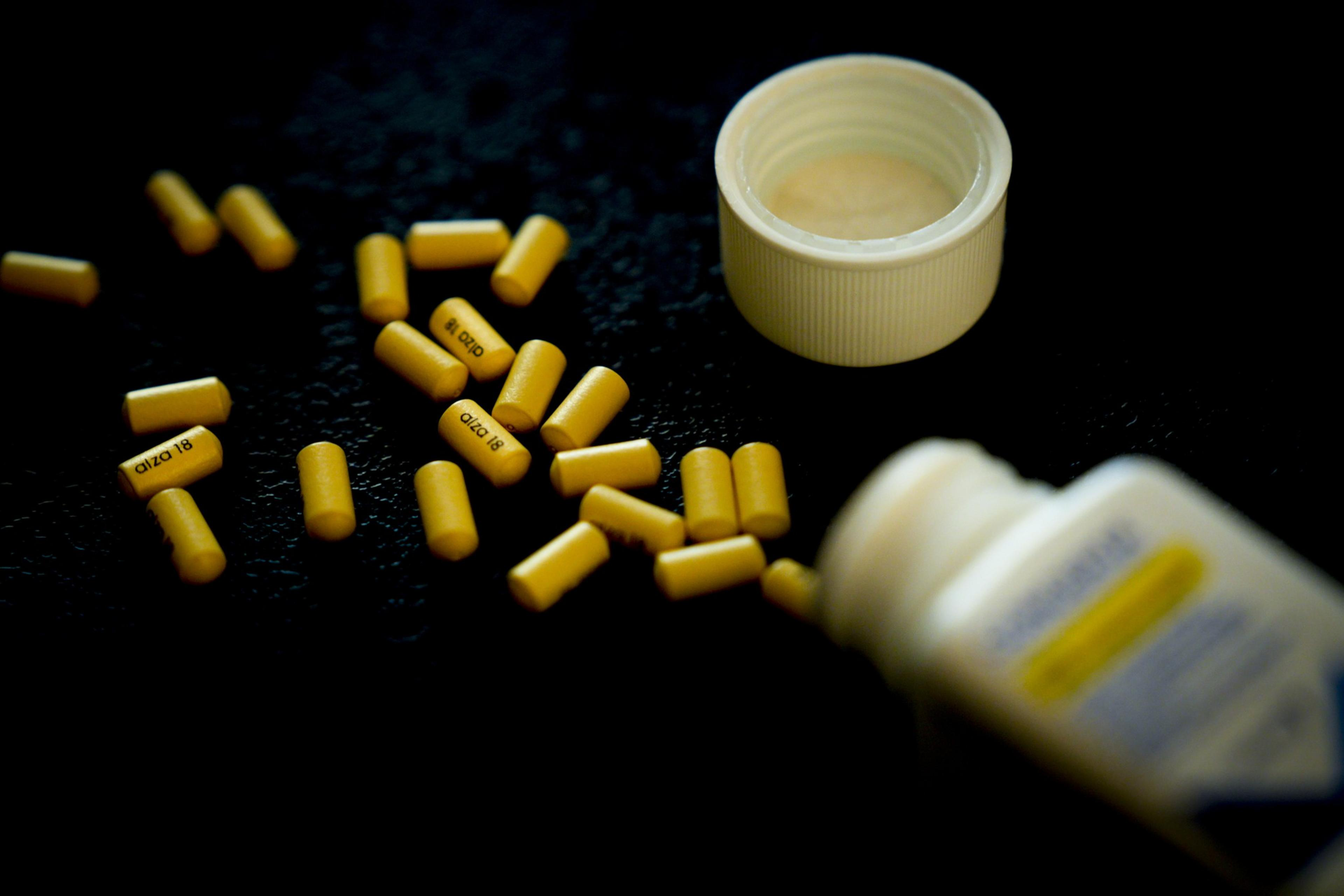 Yellow capsules have spilled onto a dark surface from a white, open pill bottle, which has its cap next to it. Some capsules are scattered around.