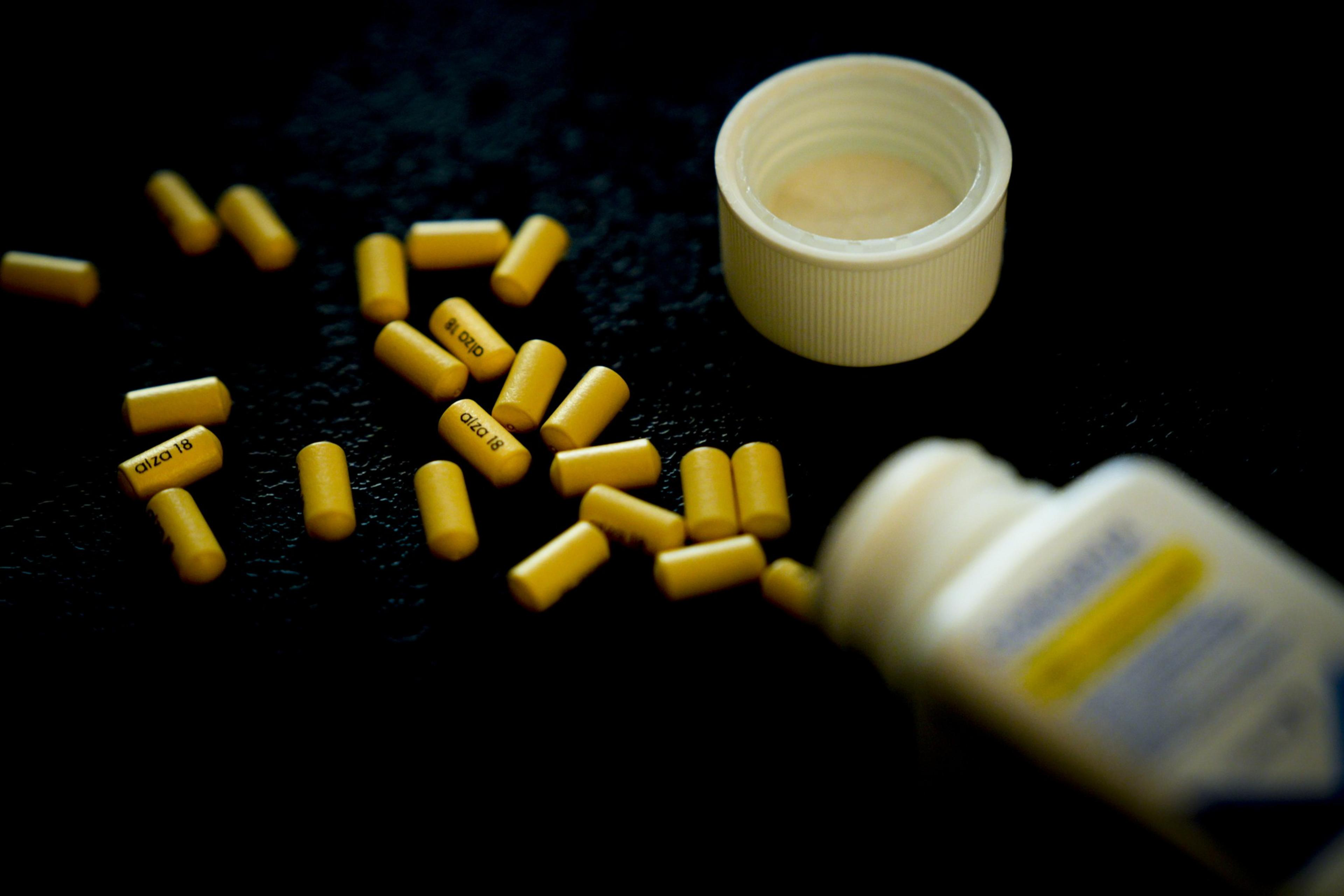 Yellow capsules have spilled onto a dark surface from a white, open pill bottle, which has its cap next to it. Some capsules are scattered around.