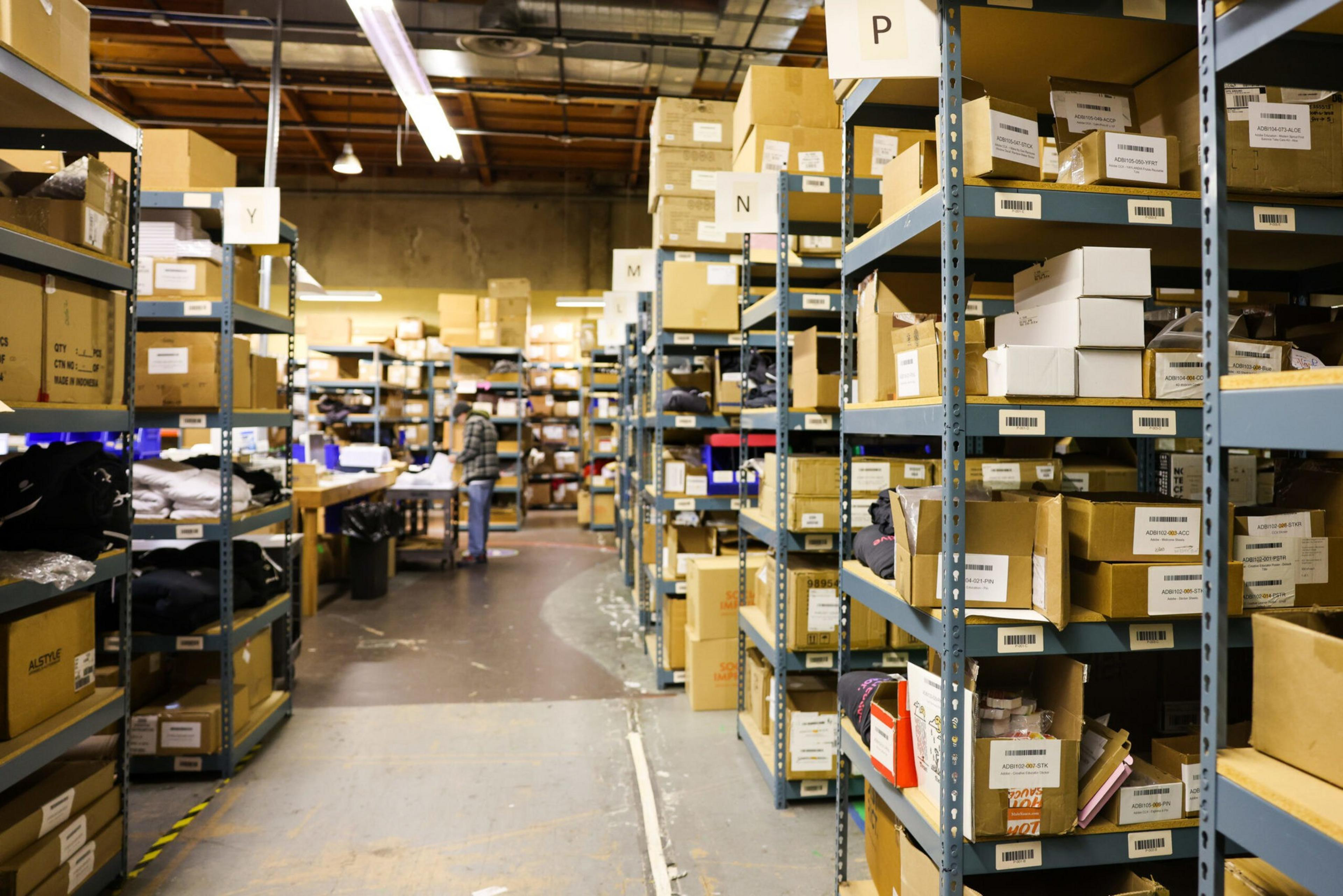 Metal shelves filled with labeled cardboard boxes line both sides of a warehouse aisle with a person working at a table in the background.