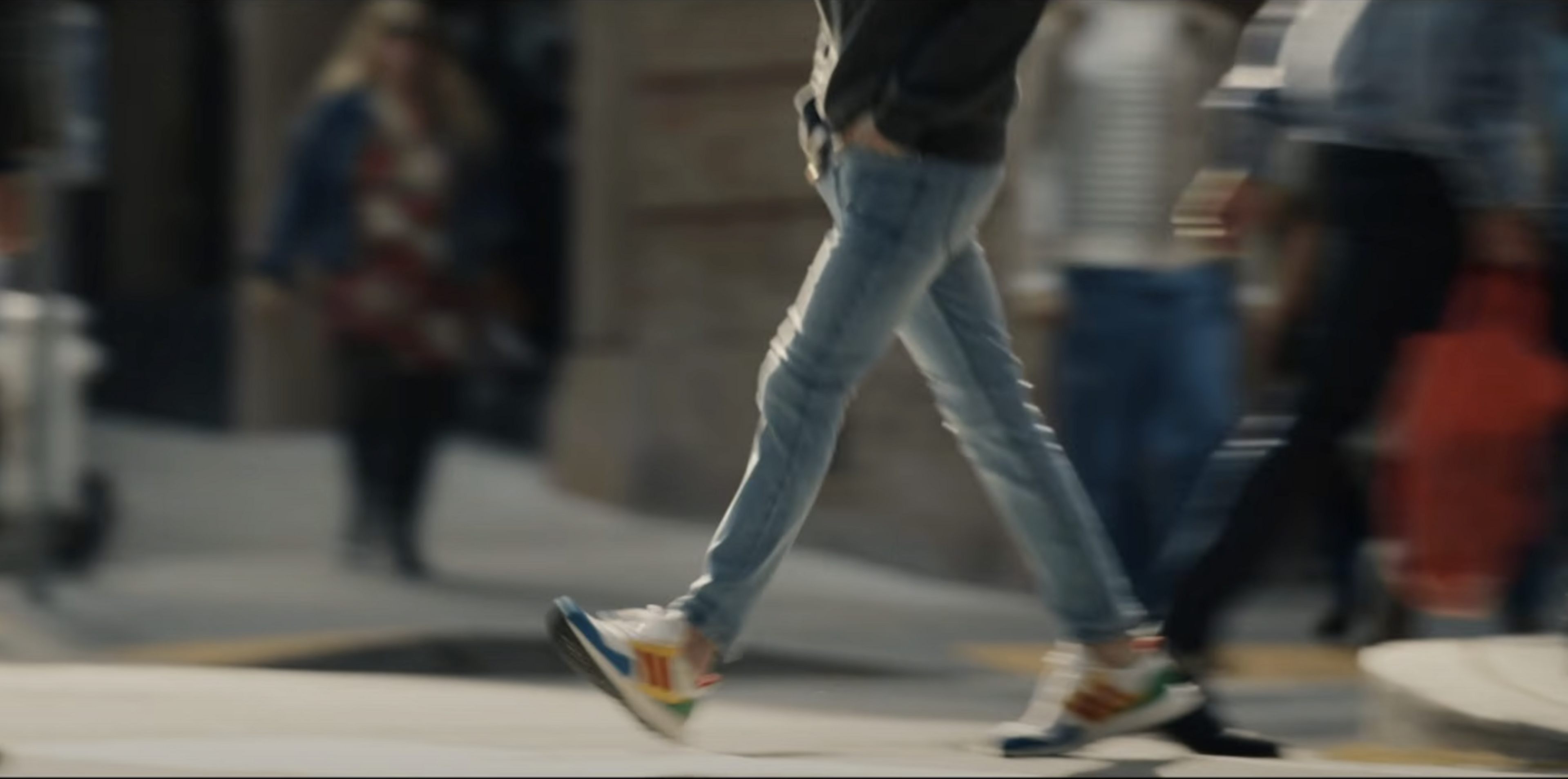 A person in jeans is walking briskly on a city street. Their hands are in their pockets, and they wear colorful sneakers. The background is blurred with people.