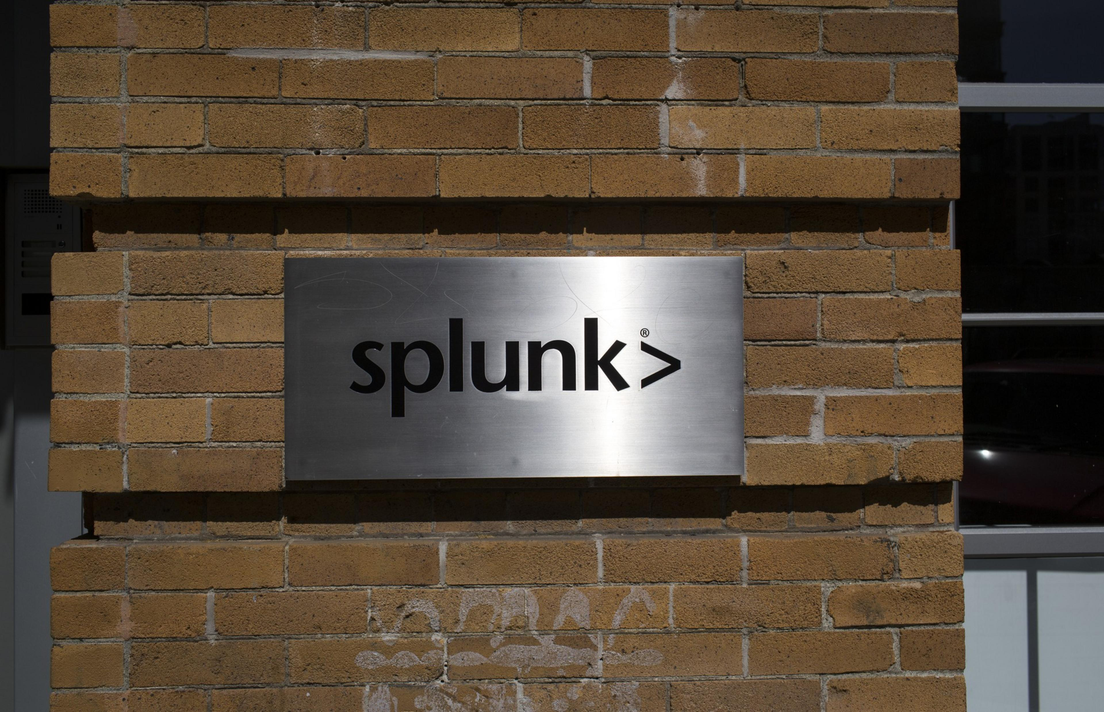 A silver-colored brushed metal sign mounted to a brick wall shines in sunlight. On the sign in lower case black lettering is a company logo.